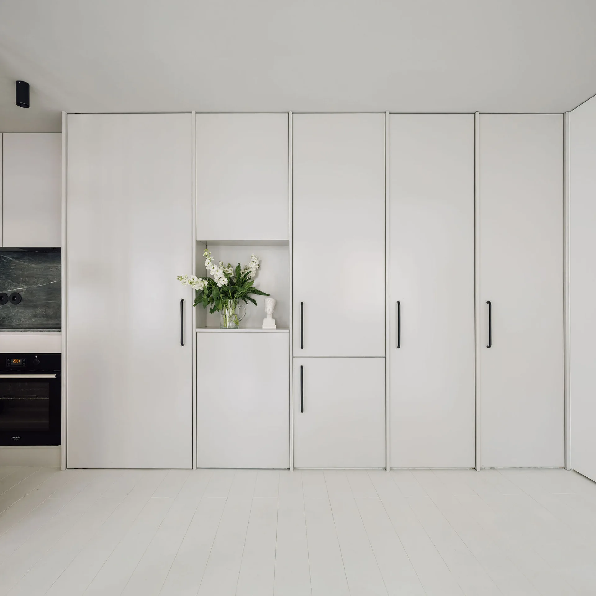 Seamless floor-to-ceiling white kitchen cabinets feature long black handles, a white plank floor, and a small open niche displaying white flowers.