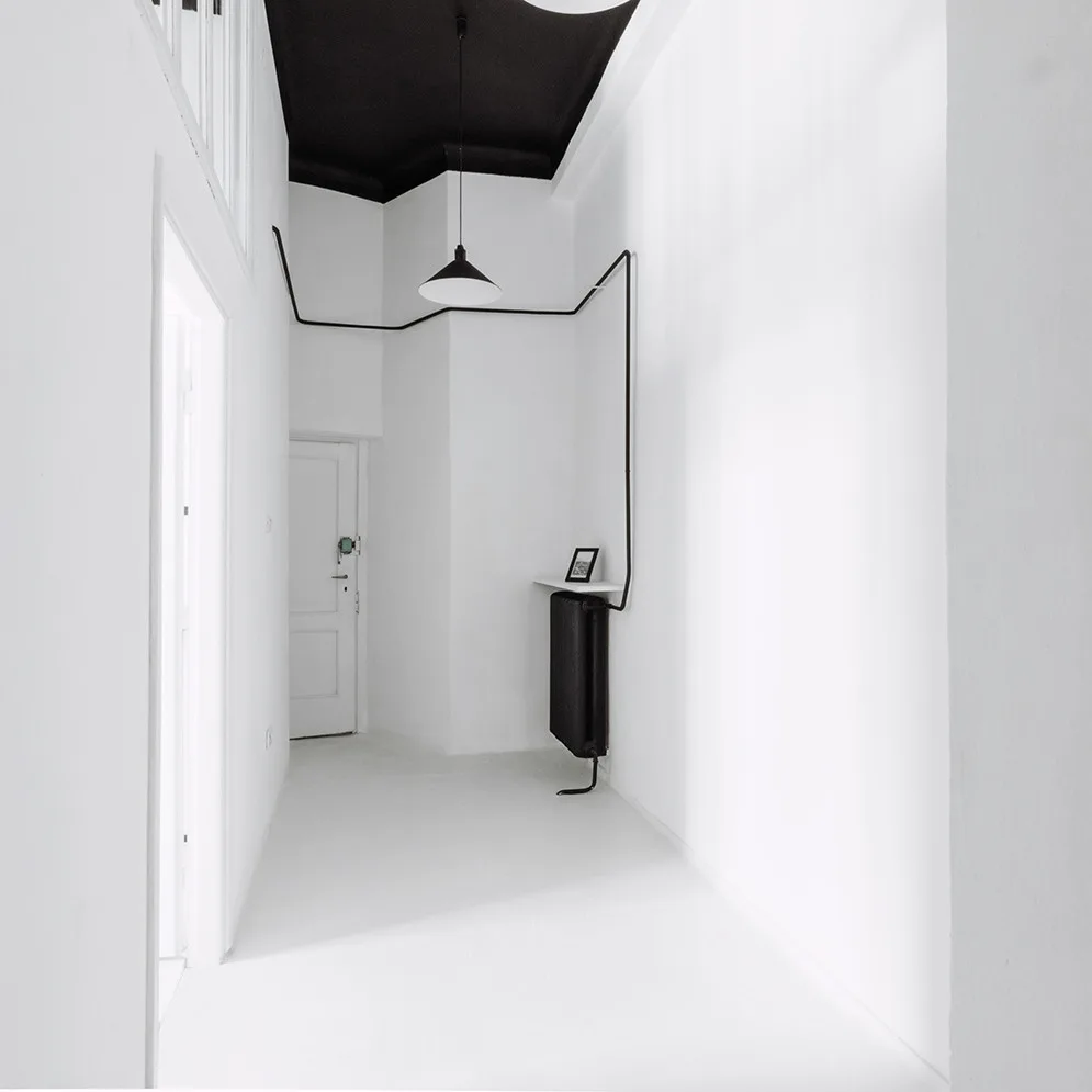 A minimalist white hallway features a stark black ceiling, a black pendant light, and a black exposed conduit pipe winding along the wall.