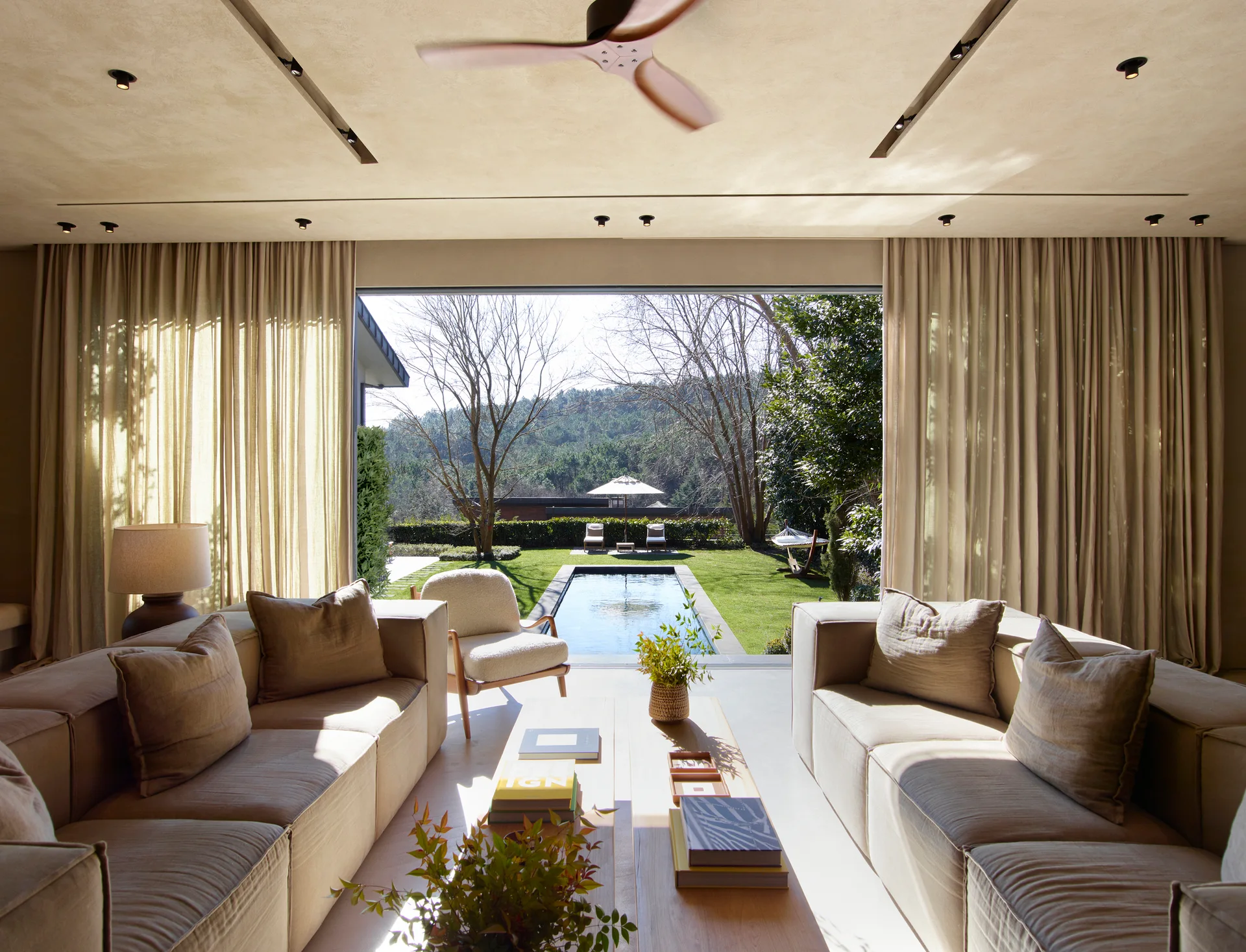 A contemporary beige living room features modular sofas and a wood coffee table facing a large opening to a backyard pool.