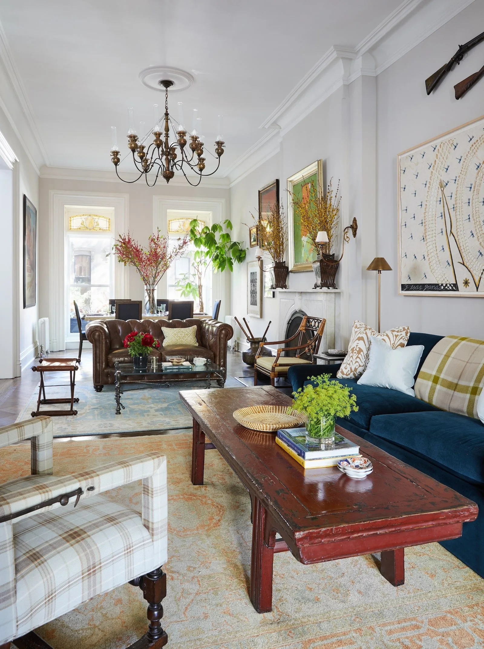 An eclectic living room featuring a long red wood coffee table, a blue sofa, a tufted brown leather sofa, and a stone fireplace.
