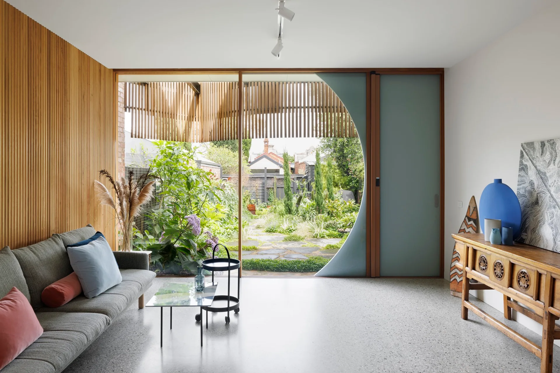 A modern living room featuring a grey sofa, a wooden console, and a large circular glass window opening to a lush garden.