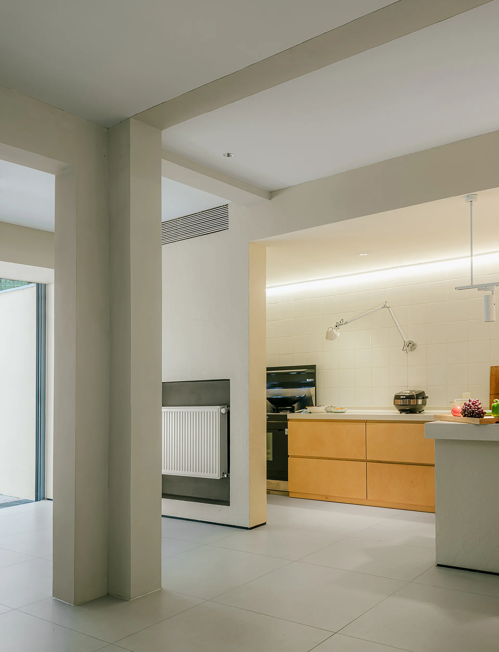 A modern minimalist kitchen features light wood cabinets, a white square-tiled backsplash, and prominent off-white structural columns.