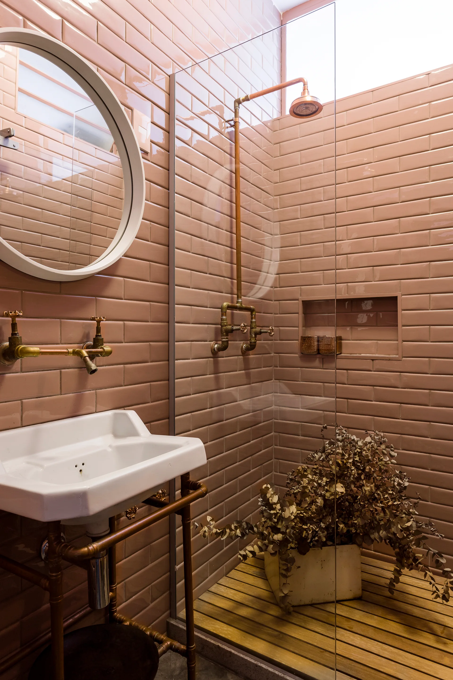 A bathroom featuring glossy pink subway tile walls, a white console sink with exposed brass plumbing, and a glass-enclosed shower.