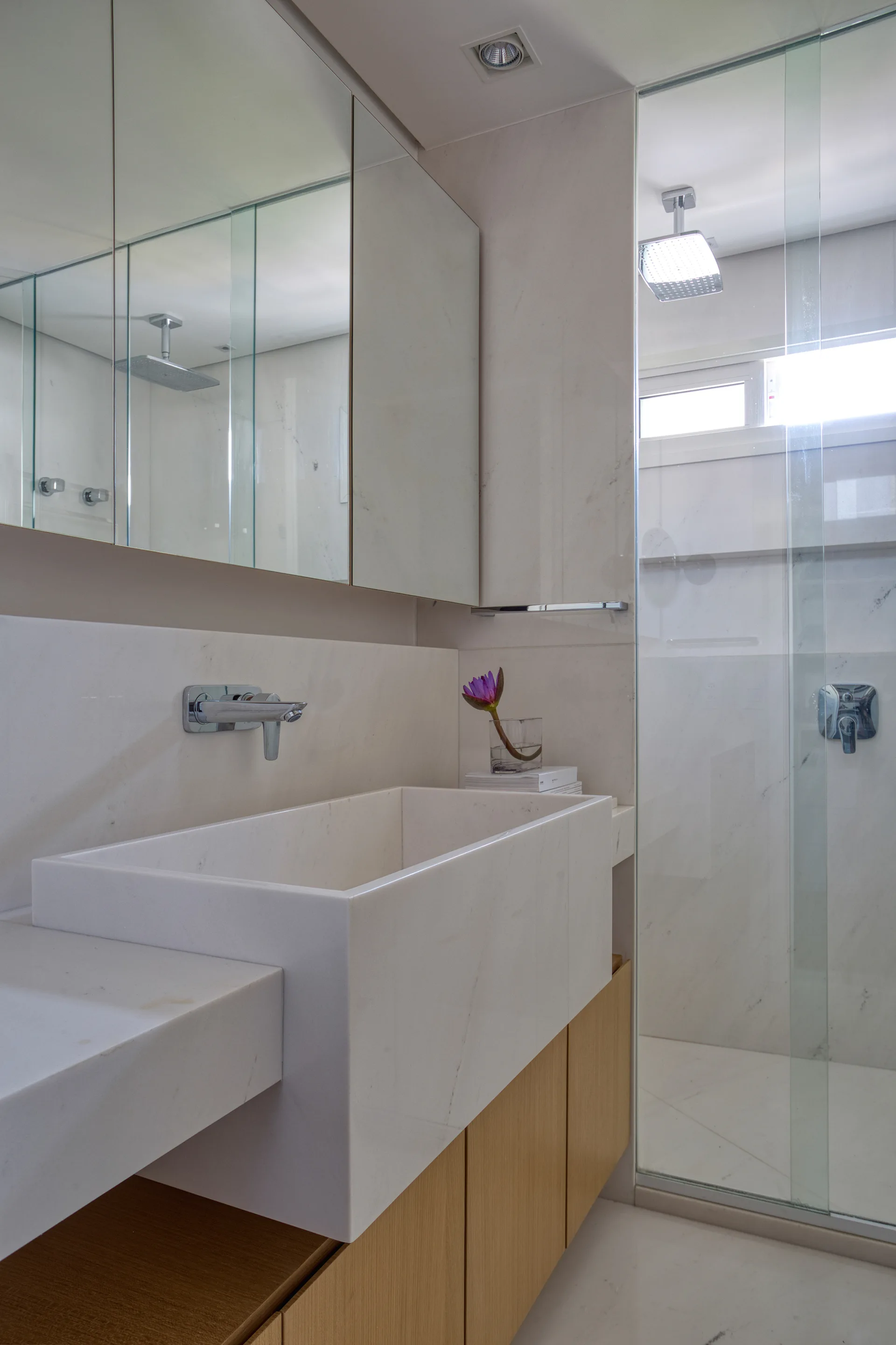 A minimalist bathroom features a white marble integrated sink over light wood cabinetry, with a glass-enclosed shower and a mirrored cabinet.