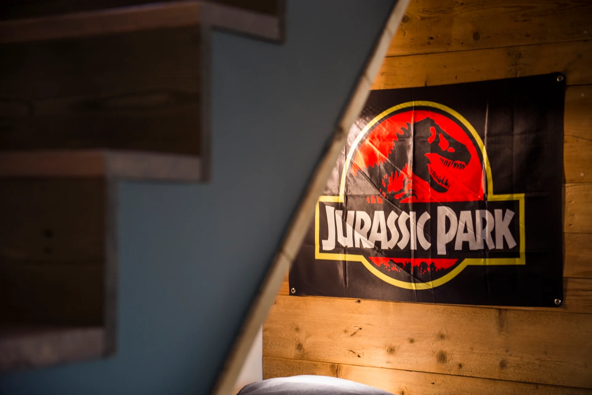 A black Jurassic Park banner hangs on a horizontal wood plank wall, partially obscured by a dark blue staircase in the foreground.