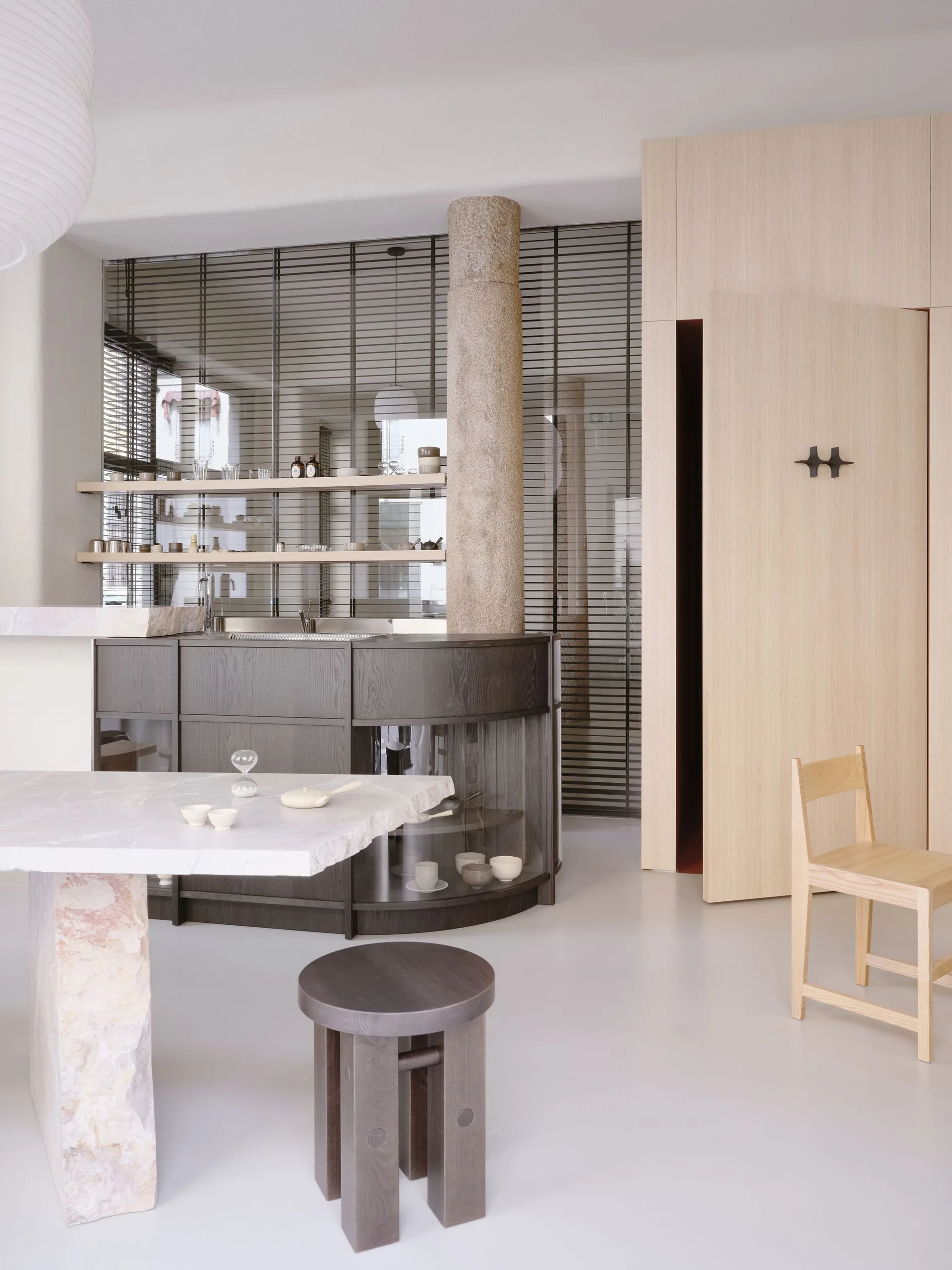 A minimalist kitchen featuring a curved dark wood island, a rough stone column, a marble slab table, and light wood wall panels.