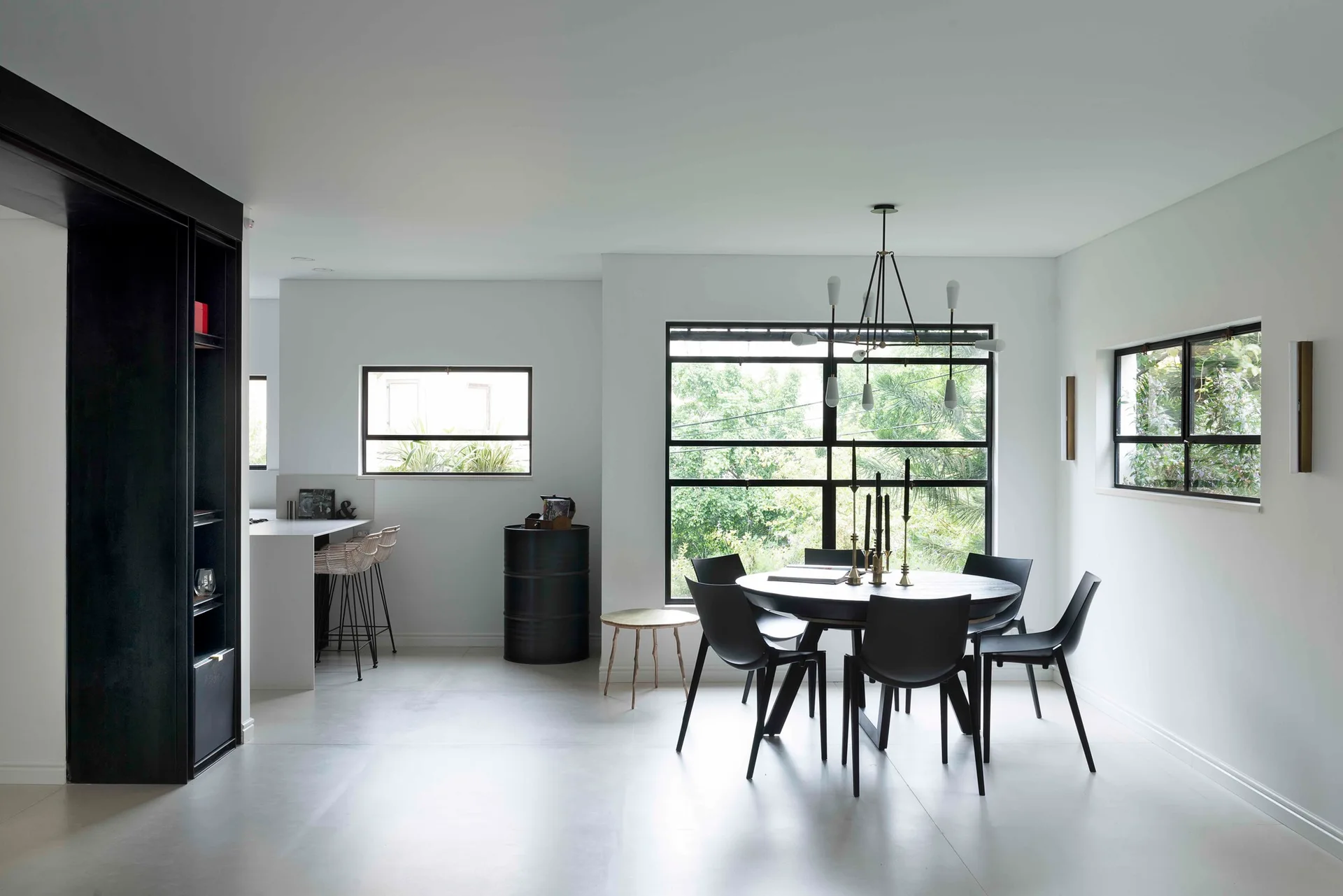 A minimalist dining area with a round black table, matching chairs, and large black-framed windows overlooking green trees.