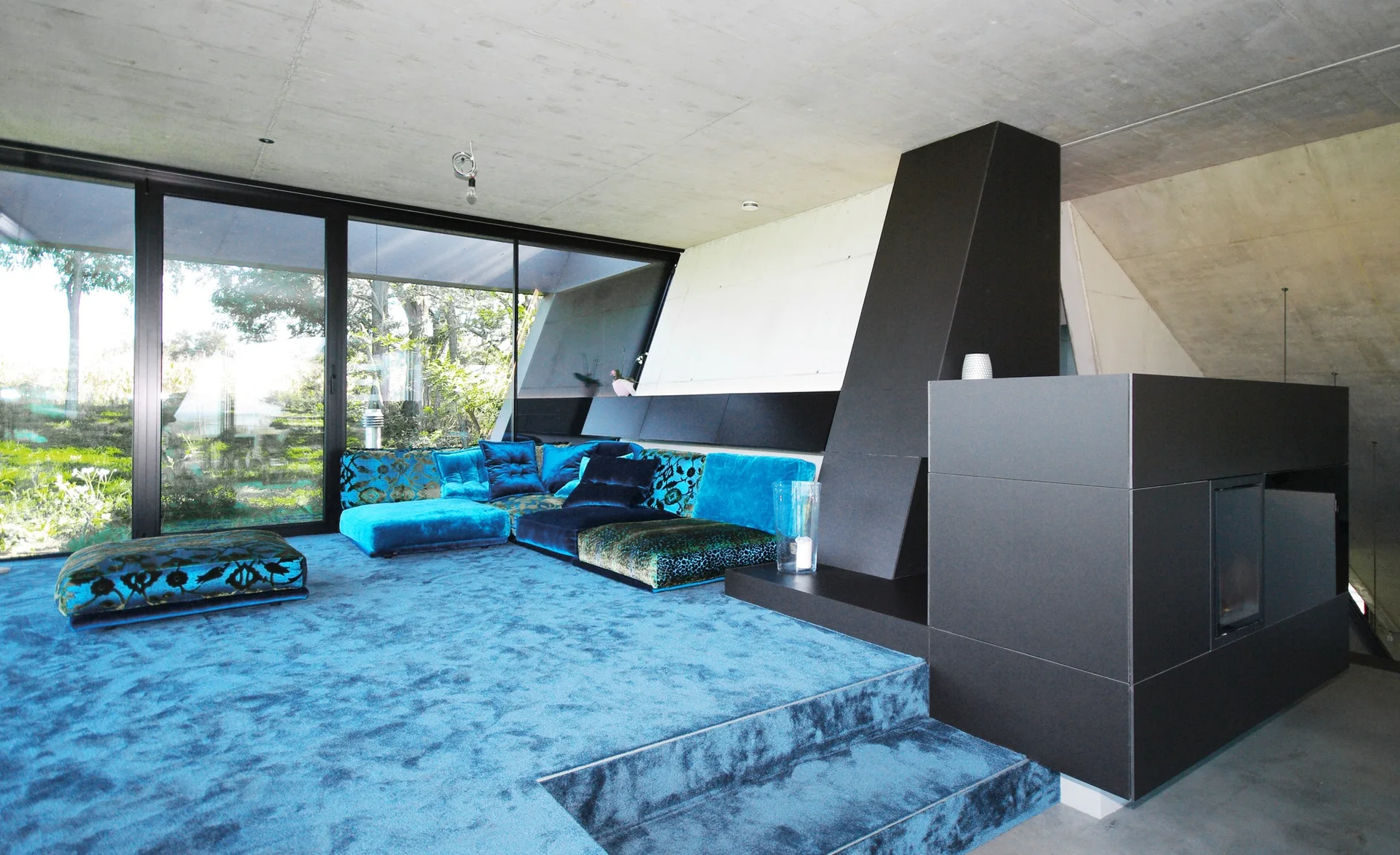 A modern living space features a blue velvet modular sofa on a raised blue carpeted platform beside a black matte fireplace and concrete ceiling.