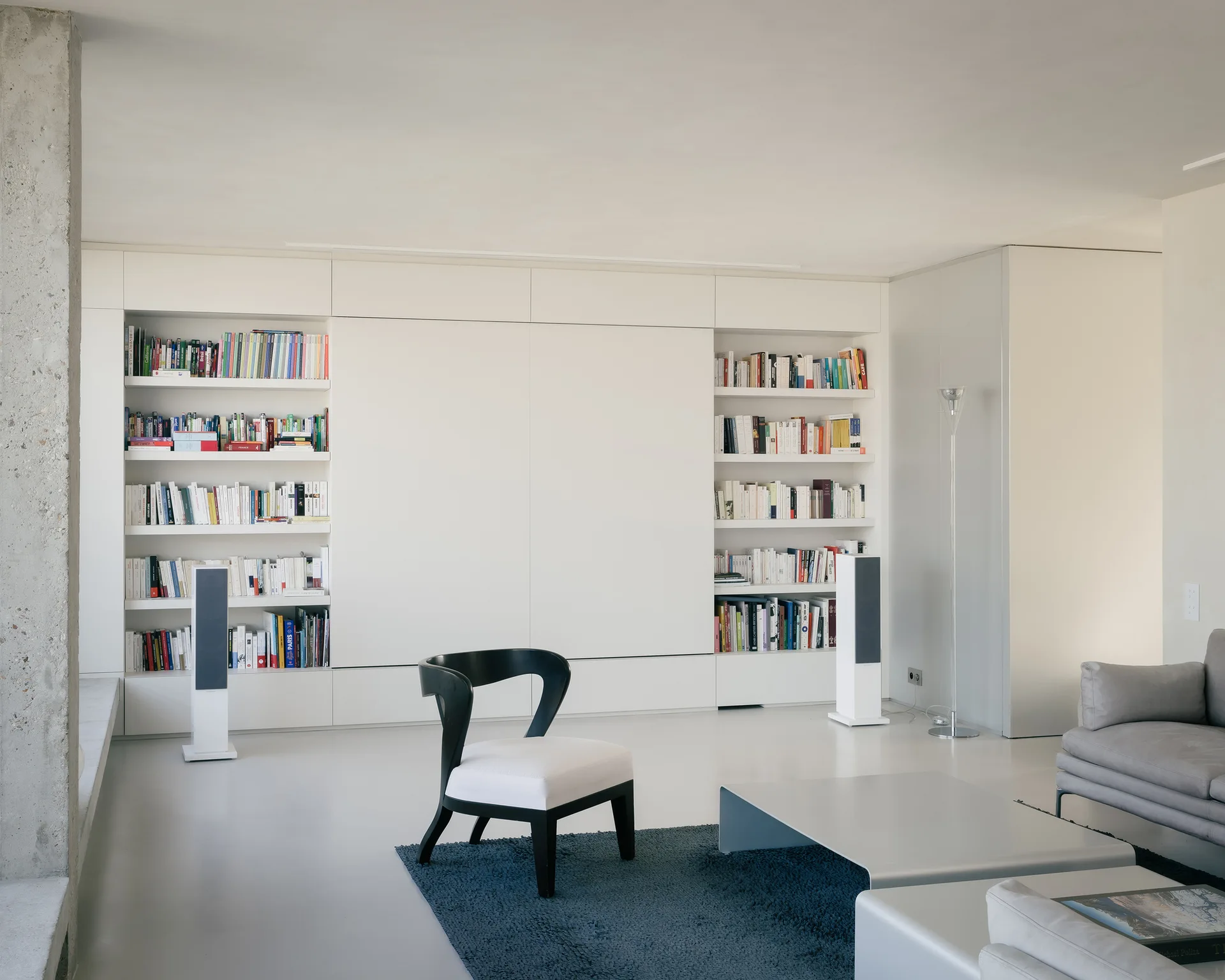 A minimalist living room featuring a large white built-in bookshelf wall, a black accent chair on a blue rug, and an exposed concrete pillar.