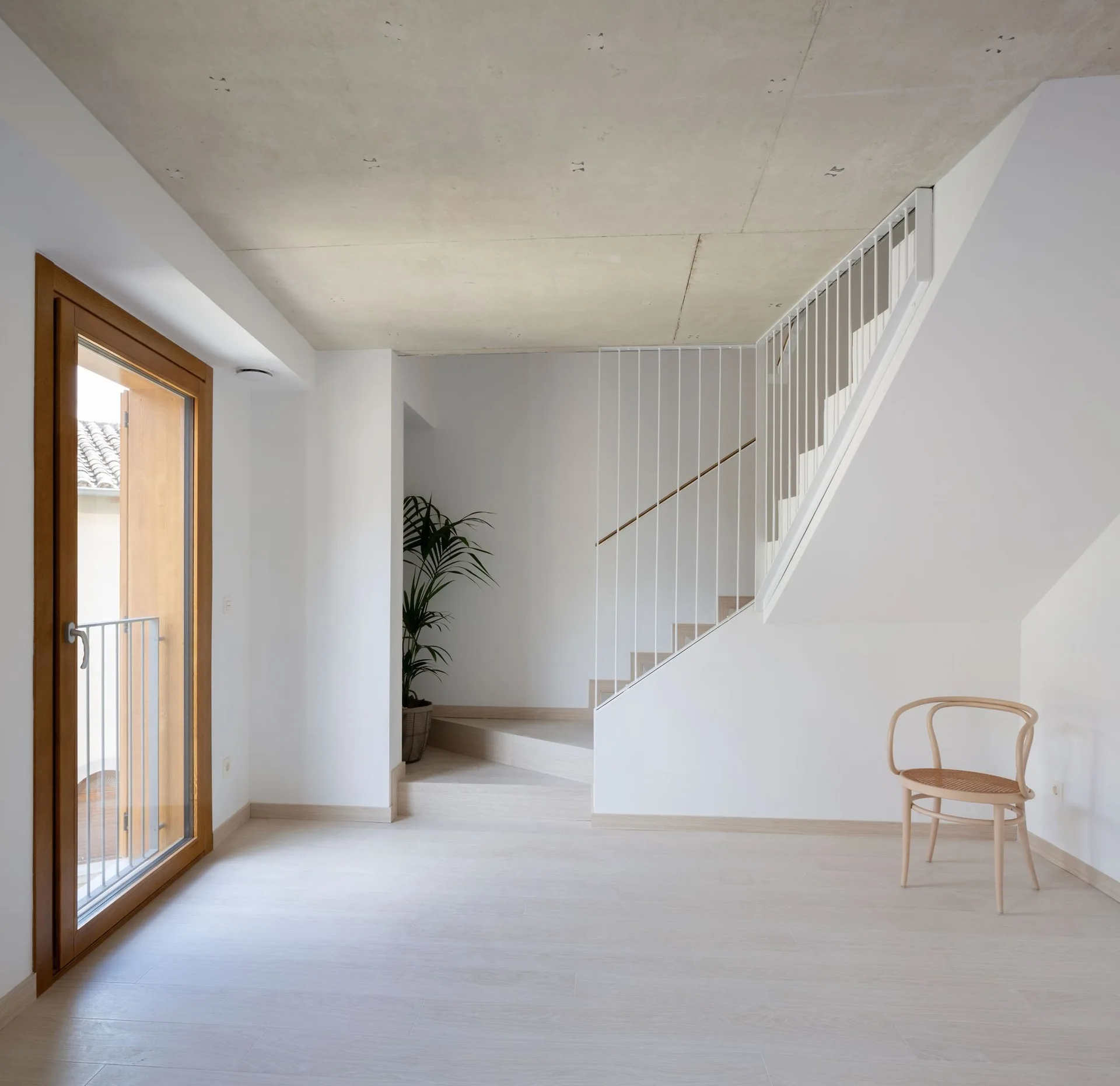 A minimalist hallway features an exposed concrete ceiling, a light wood staircase with white vertical metal railings, and a single wooden bentwood chair.