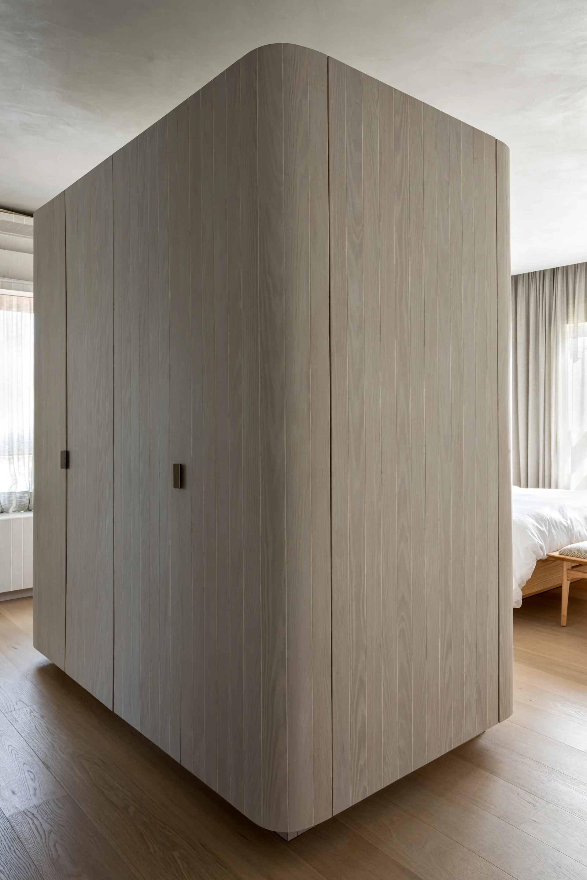 A freestanding light wood closet with rounded corners stands on a hardwood floor, with a bed and curtains visible in the background.