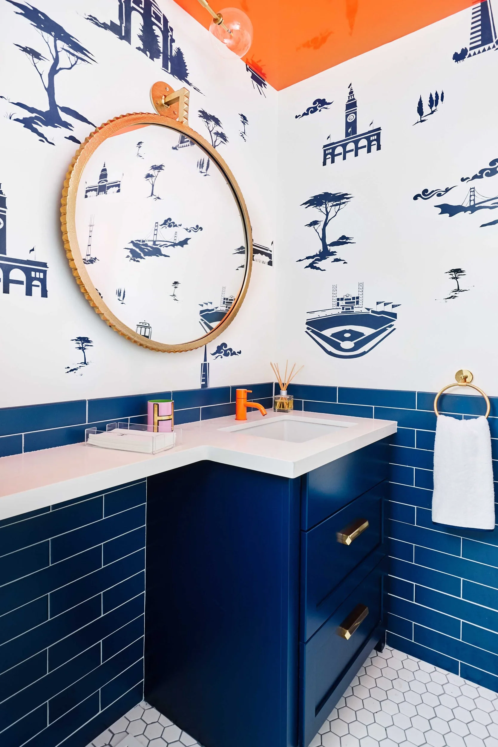 A small bathroom features blue subway tile wainscoting, illustrative blue and white wallpaper, a blue vanity, and a round gold mirror.