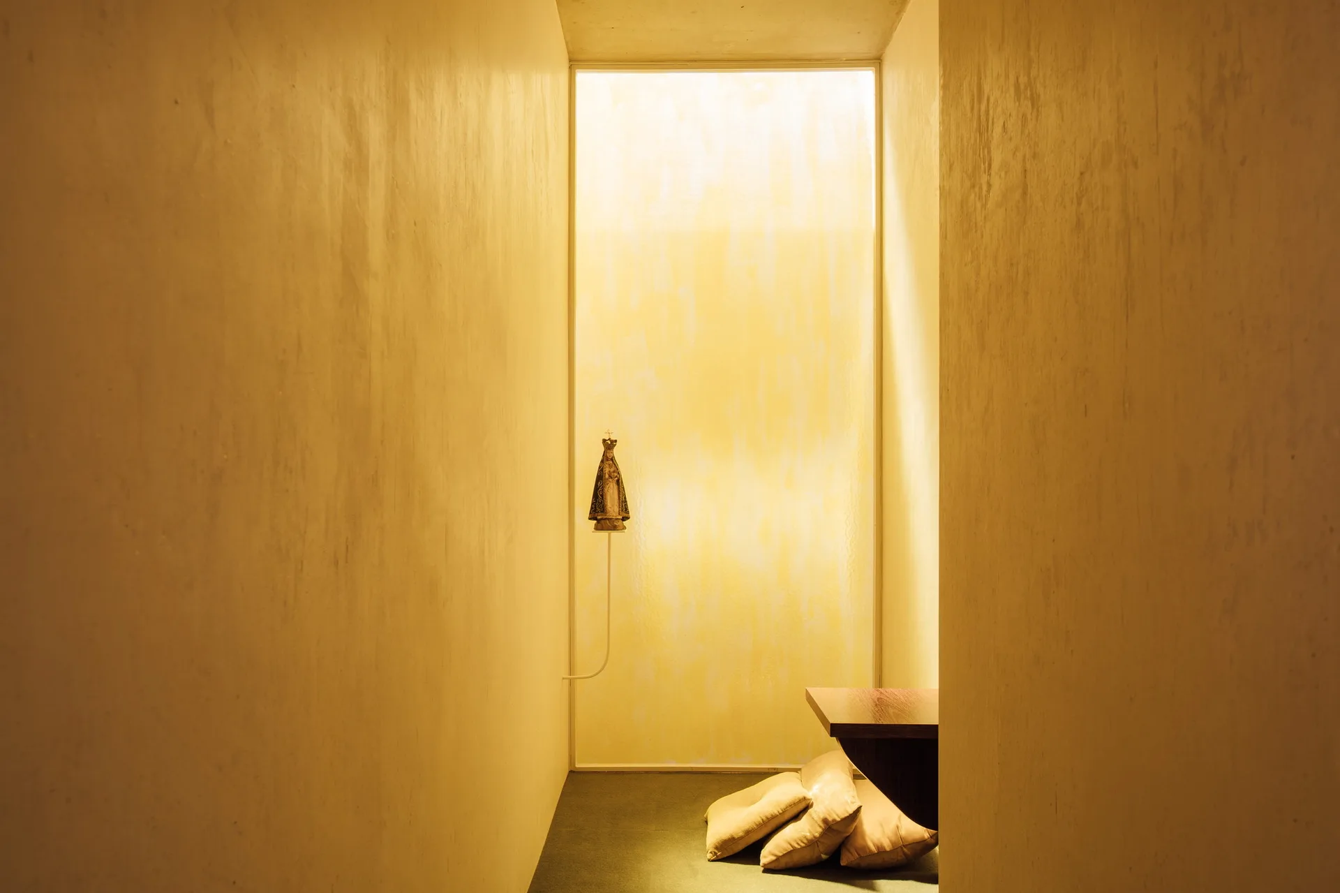 A minimalist yellow hallway with a large backlit translucent window, a floating dark wood shelf, floor cushions, and a slender wall bracket.