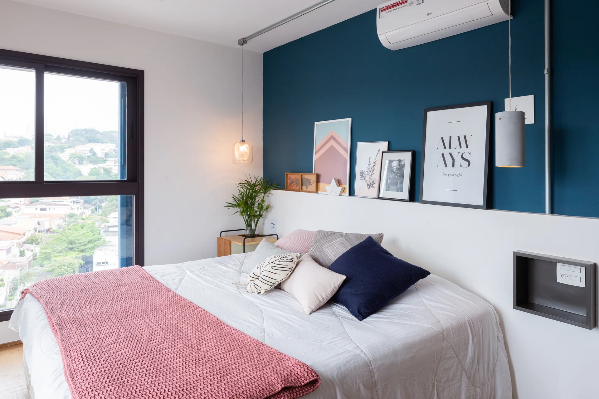 A modern bedroom features a bed with pink and blue pillows, a teal accent wall with a white shelf displaying art frames, and a large window.