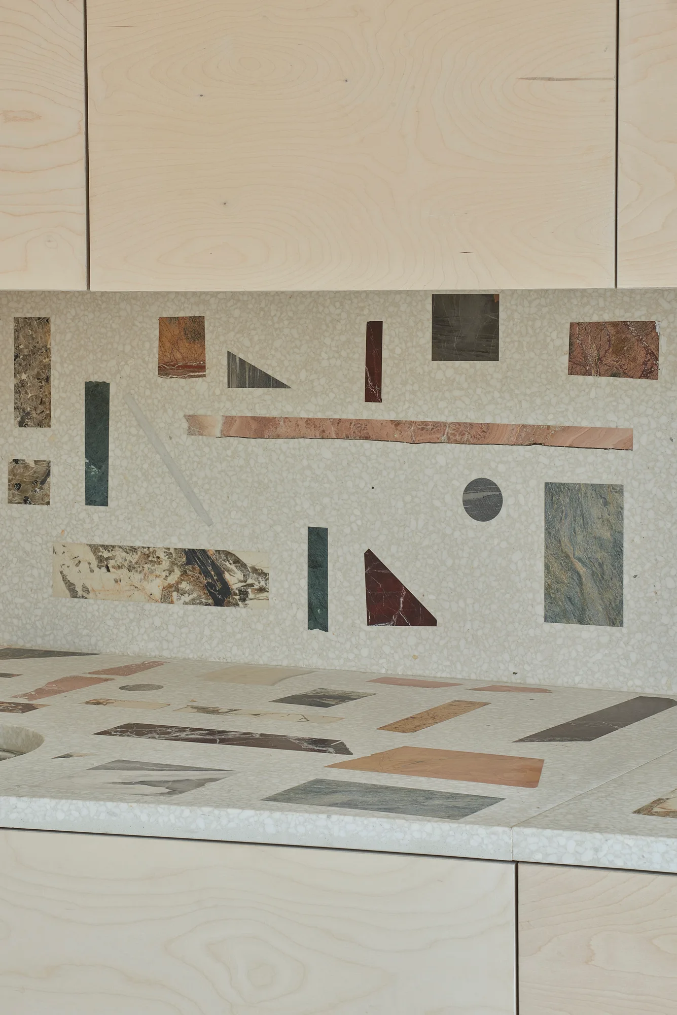 A close-up of a neutral terrazzo kitchen countertop and backsplash inlaid with multi-colored geometric marble shapes beneath light wood cabinets.