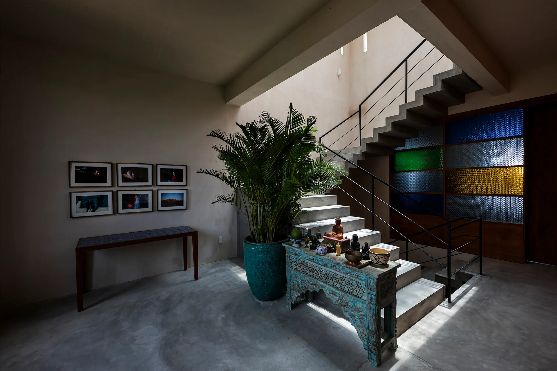 A concrete floating staircase ascends next to a carved turquoise console table, a large potted palm, and a multicolored stained glass wall.