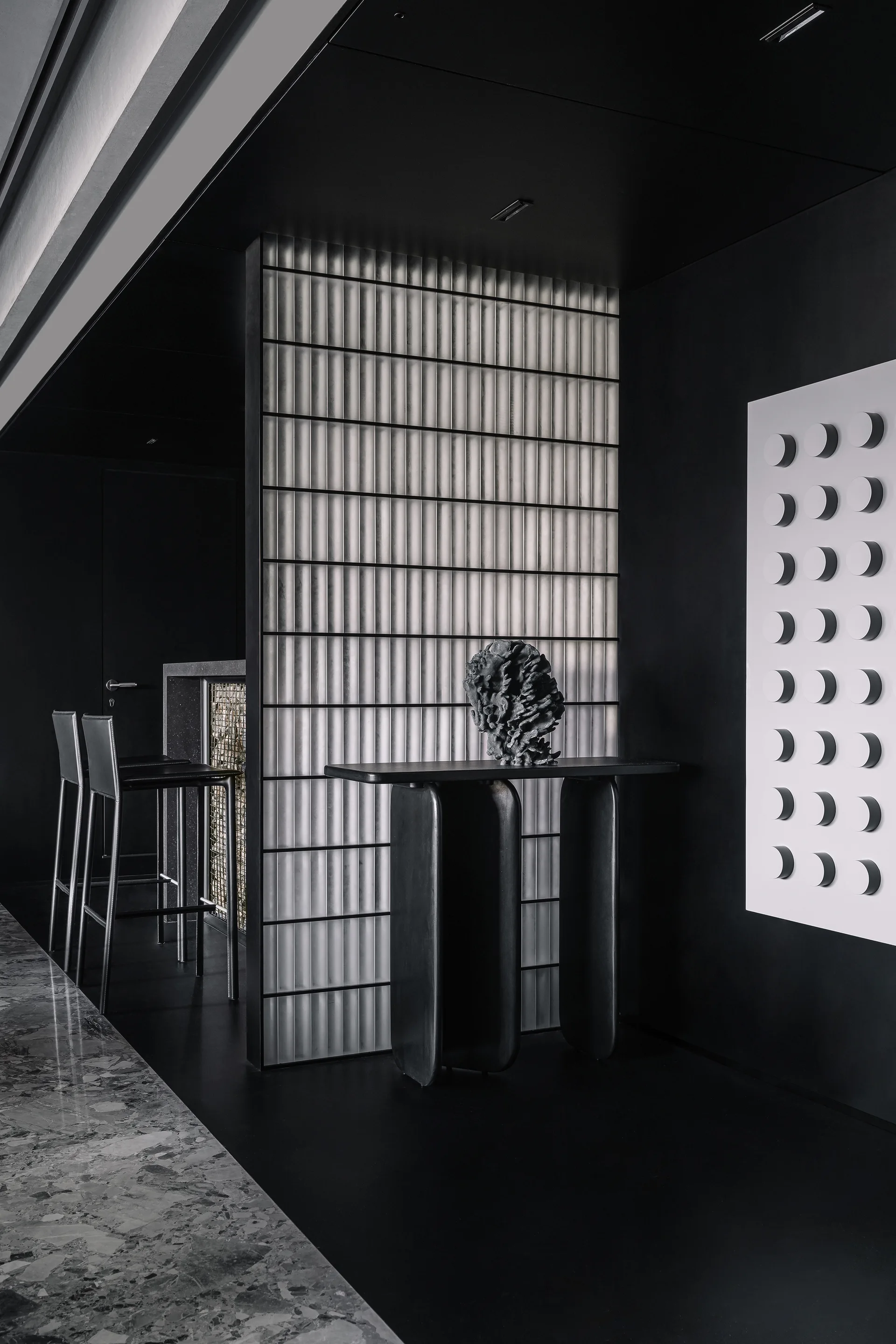 A dark modern lounge features a fluted glass partition behind a black console table, with two bar stools visible in the background.