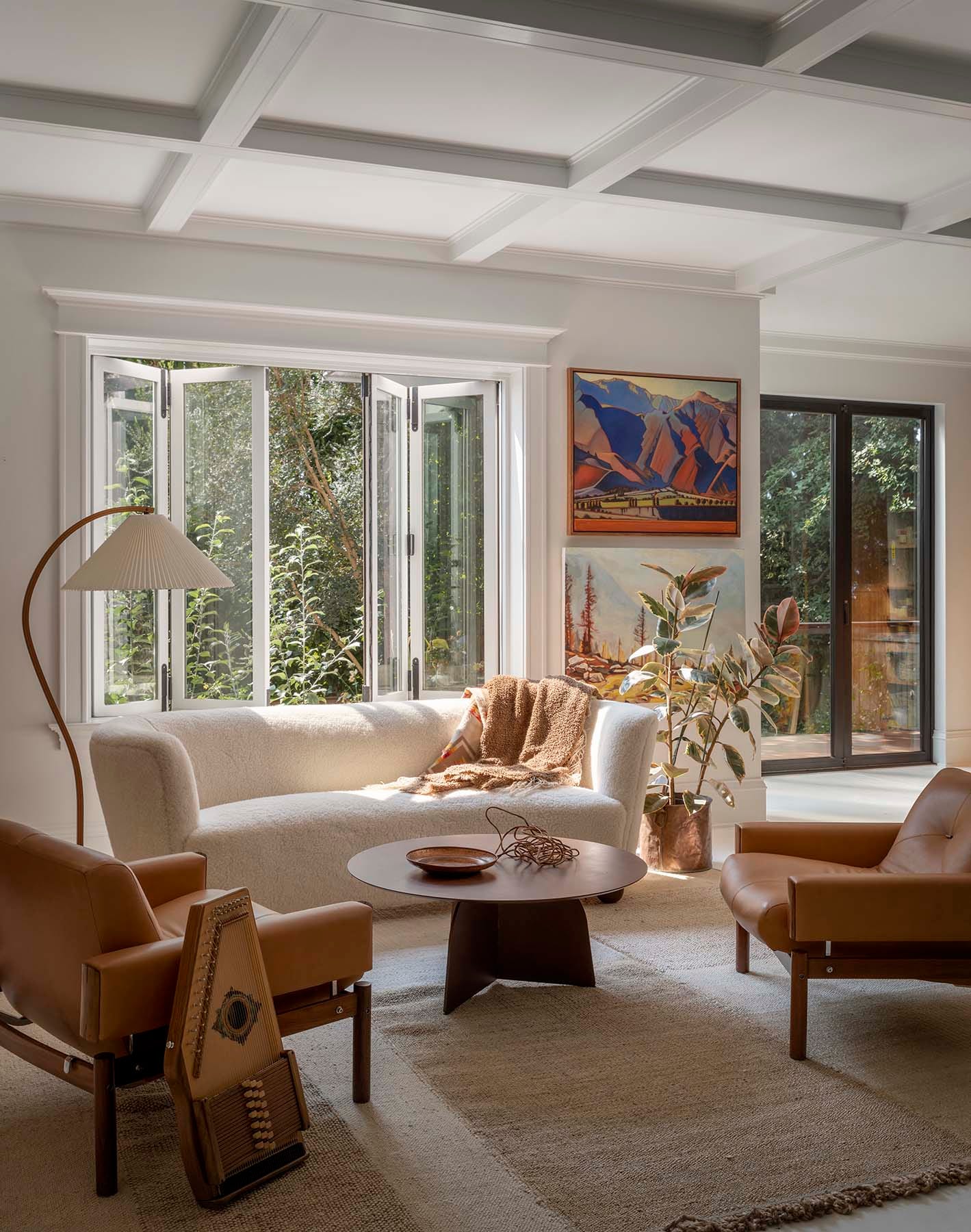 A bright living room features a curved white bouclé sofa, tan leather armchairs, a wooden coffee table, and a white coffered ceiling.