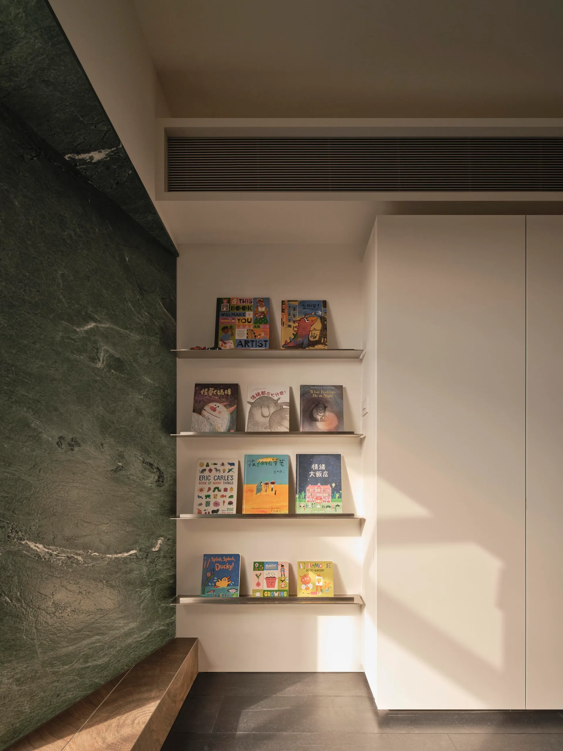 A recessed white wall niche with metal floating shelves displaying books next to a textured green stone wall and a white storage cabinet.