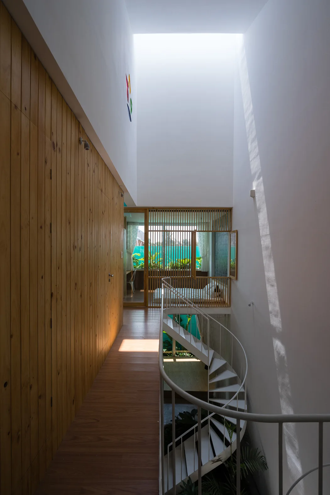 A white spiral staircase winds downward beneath a large rectangular skylight in a modern hallway lined with vertical wood paneling.