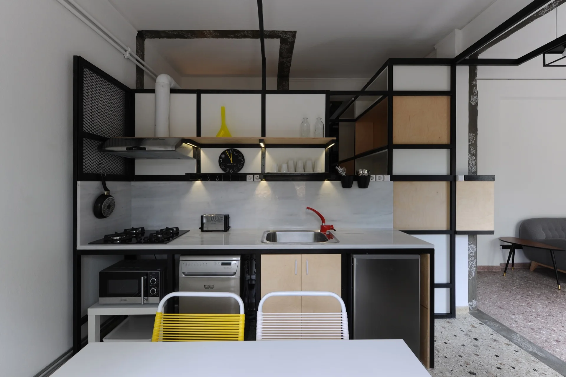 An industrial kitchen featuring a black metal frame structure, plywood cabinetry, terrazzo flooring, and two modern dining chairs in the foreground.