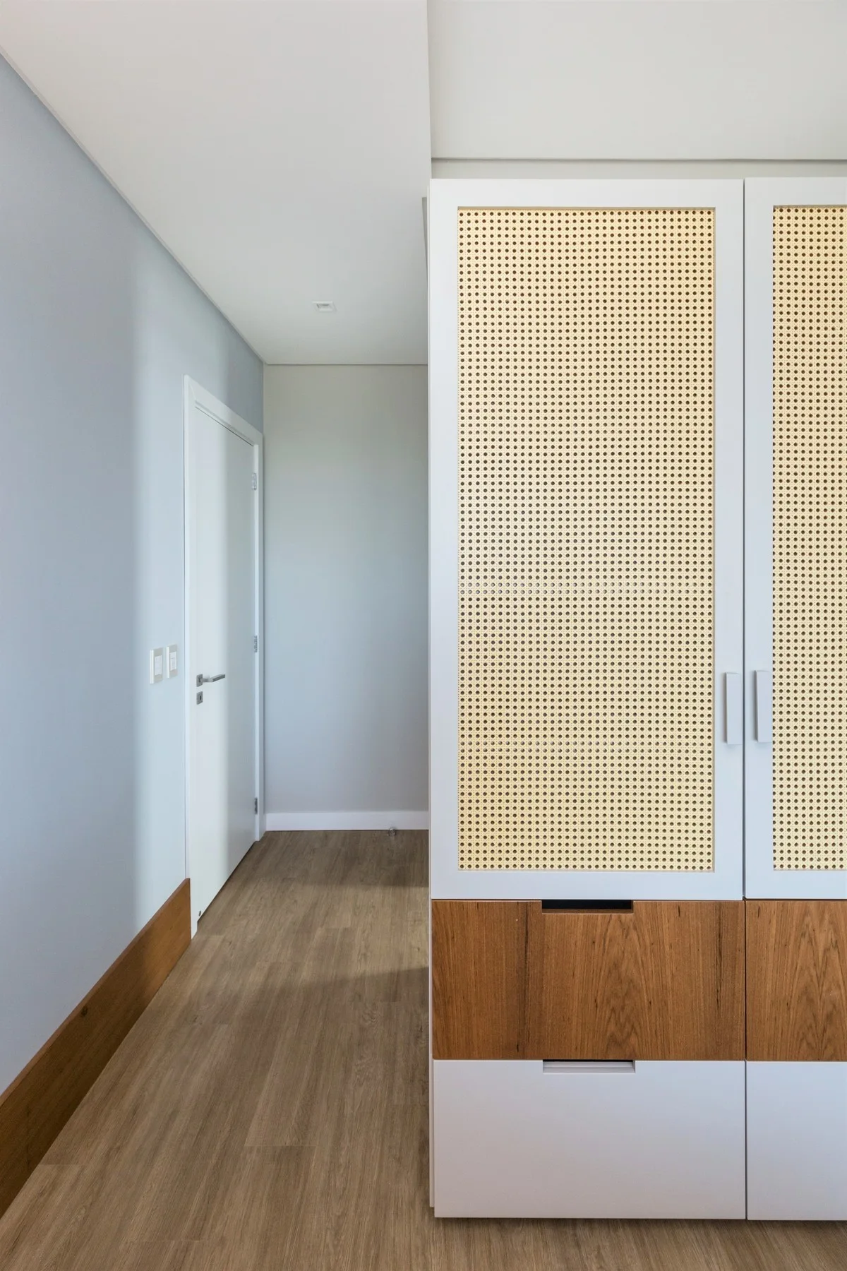 A custom white cabinet featuring woven rattan doors and wood drawers stands in a hallway with light blue walls and wood flooring.