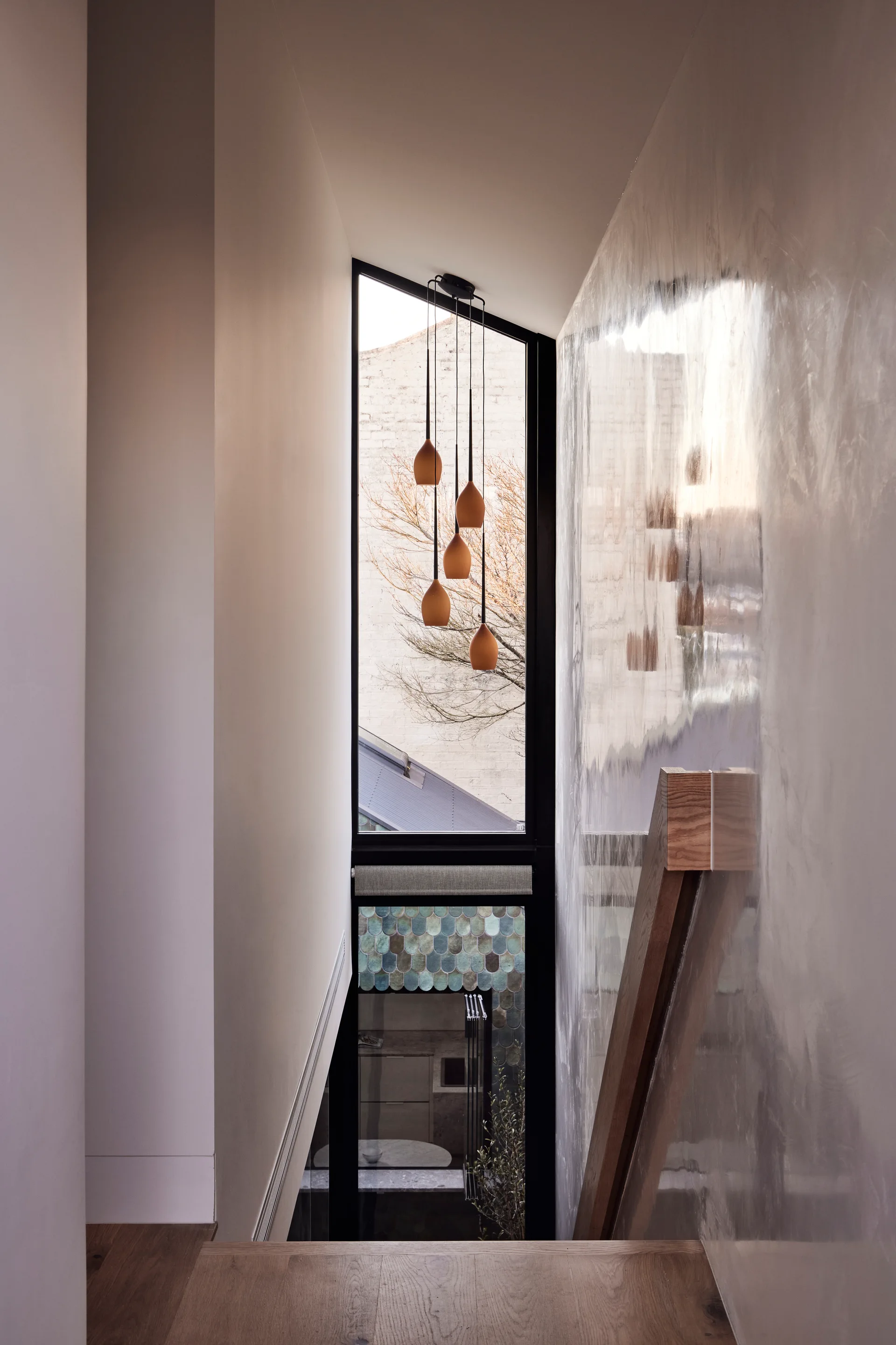 A view looking down a modern staircase featuring a reflective polished plaster wall, wooden handrail, and terracotta pendant lights in front of a tall window.