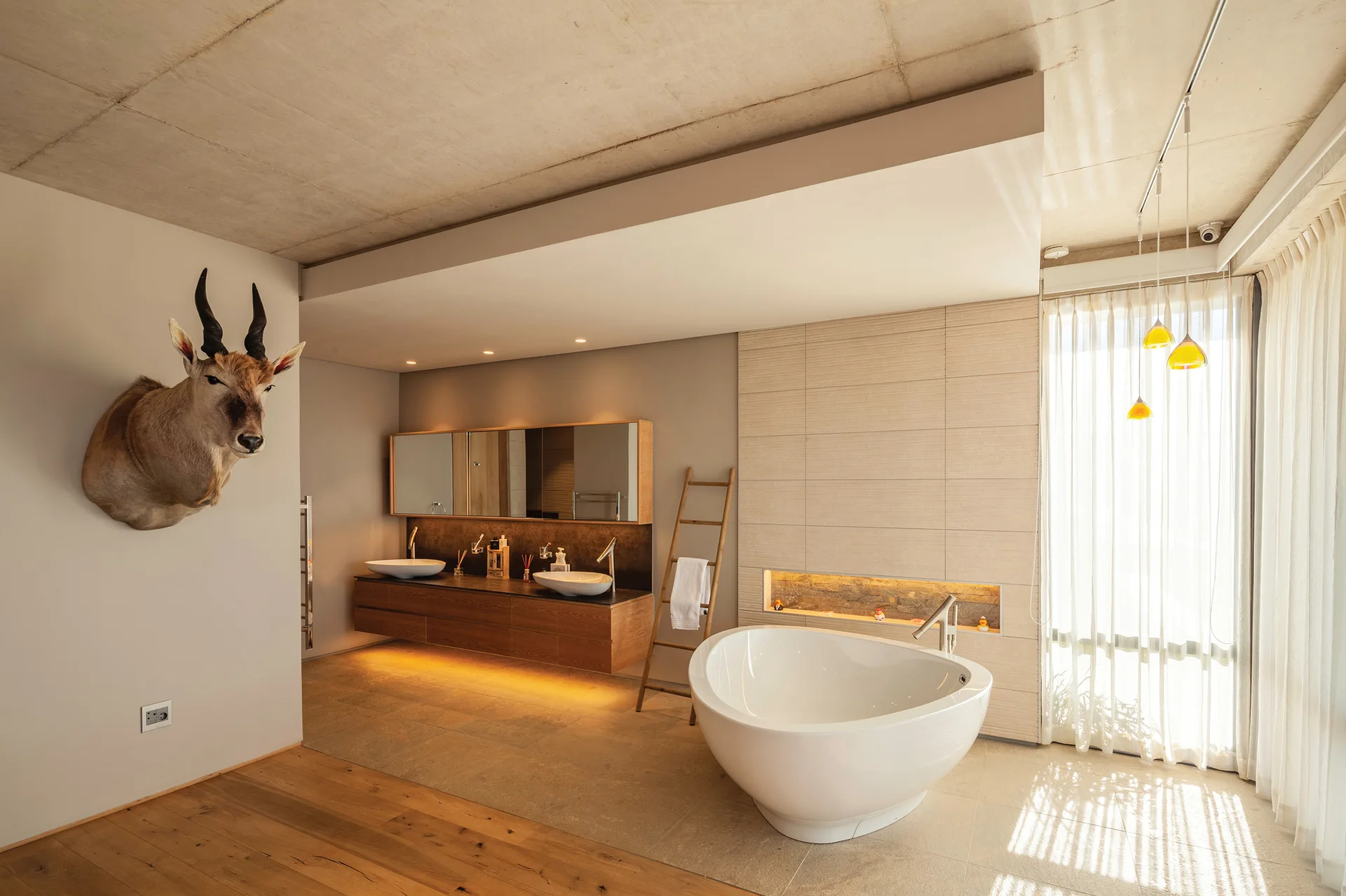 A spacious bathroom features a freestanding white soaking tub, a floating wooden vanity with two sinks, and an exposed concrete ceiling.