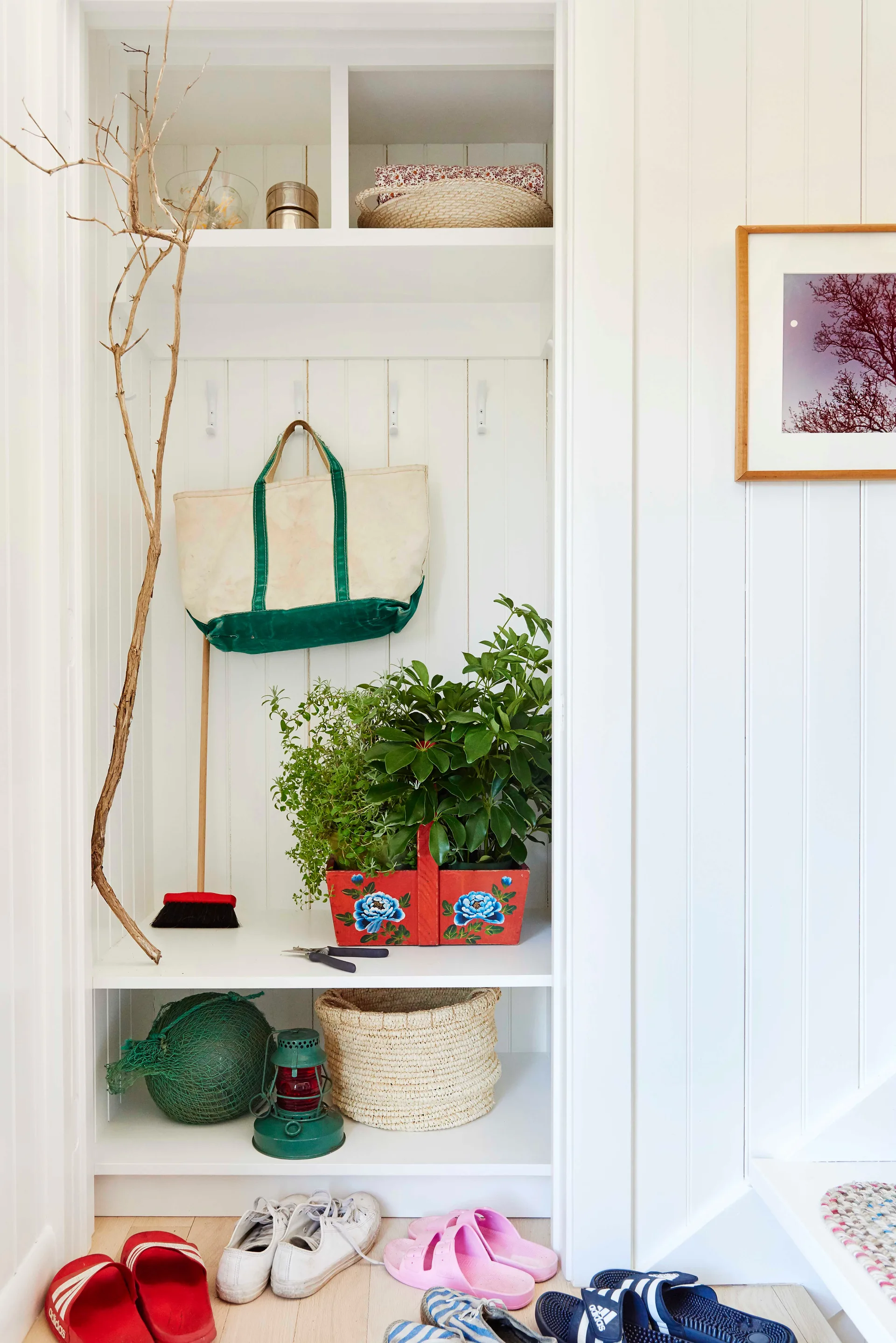 A white painted wooden mudroom nook features built-in shelves, coat hooks, a canvas tote bag, woven baskets, and potted plants.