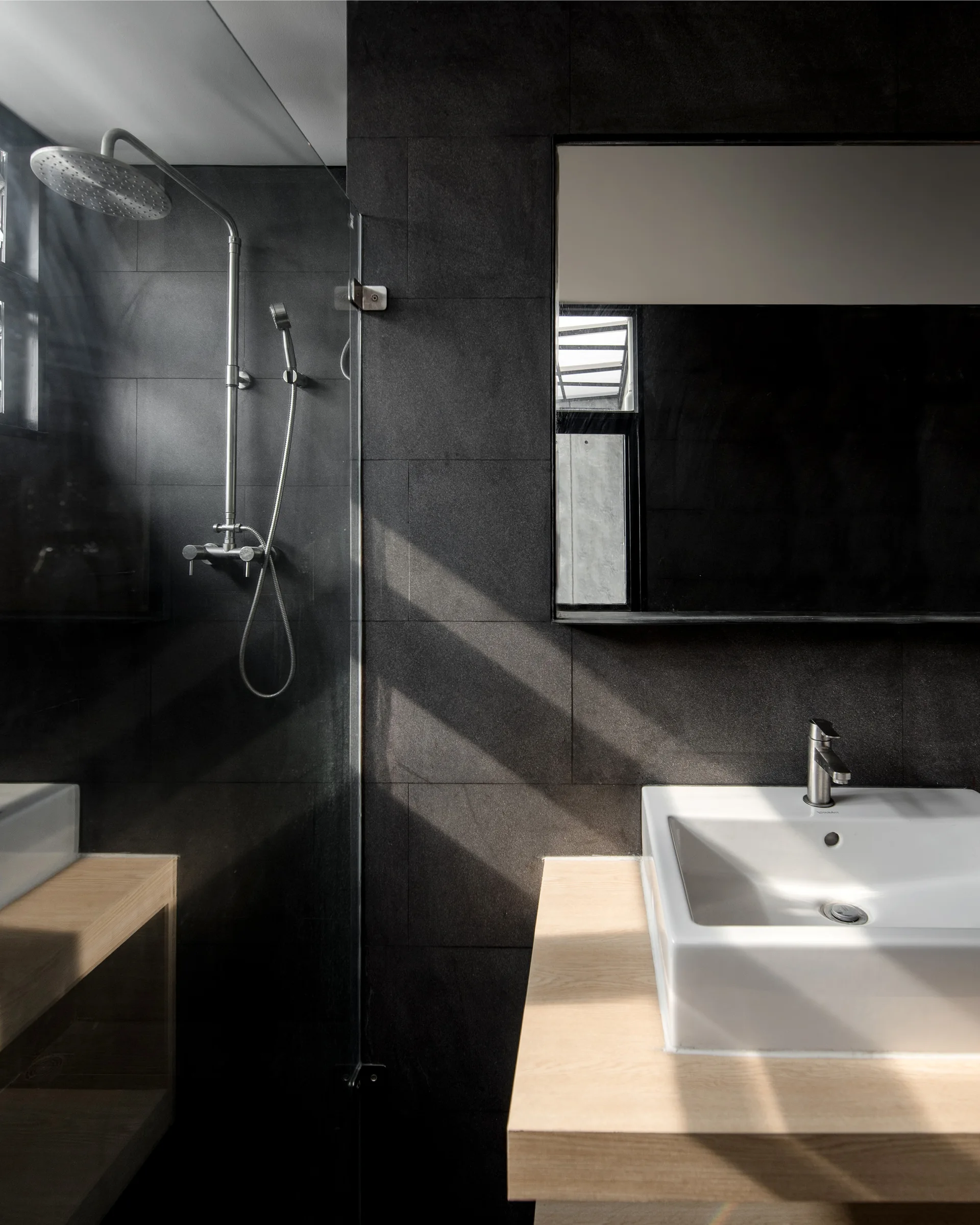 A modern bathroom features dark charcoal tiled walls, a glass shower panel with chrome fixtures, a light wood vanity, and a white ceramic sink.