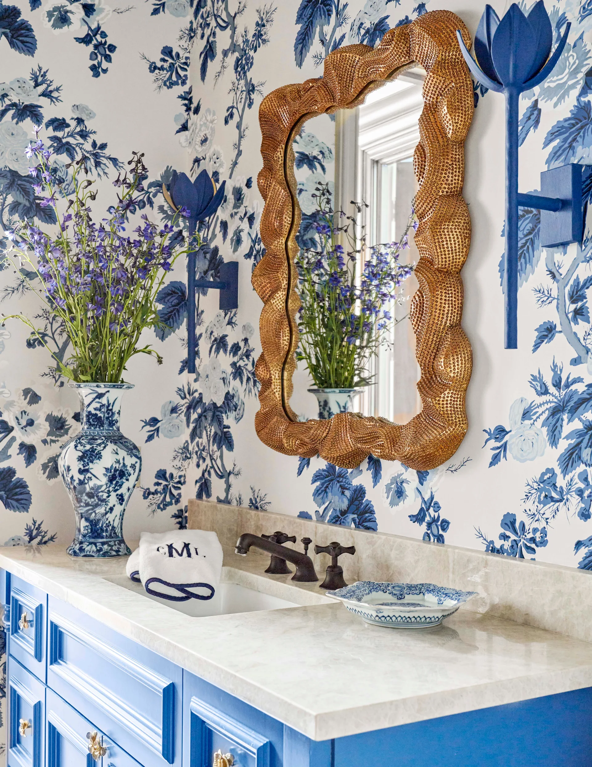 A blue bathroom vanity with a light stone countertop is paired with blue and white floral wallpaper and a textured gold wavy mirror.