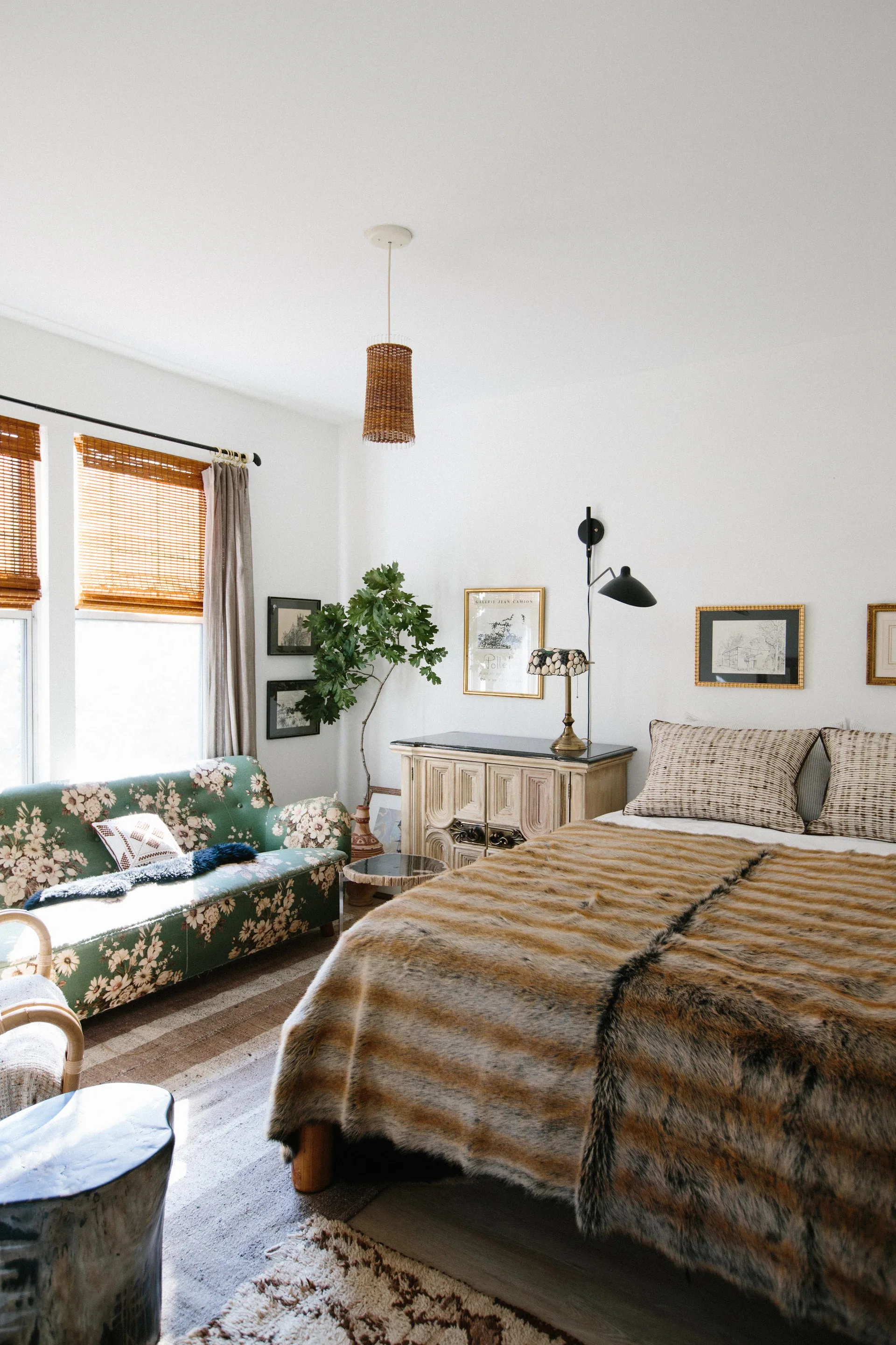 An eclectic bedroom features a green floral sofa by the window, a wood cabinet, and a bed with striped pillows and a brown faux fur throw.