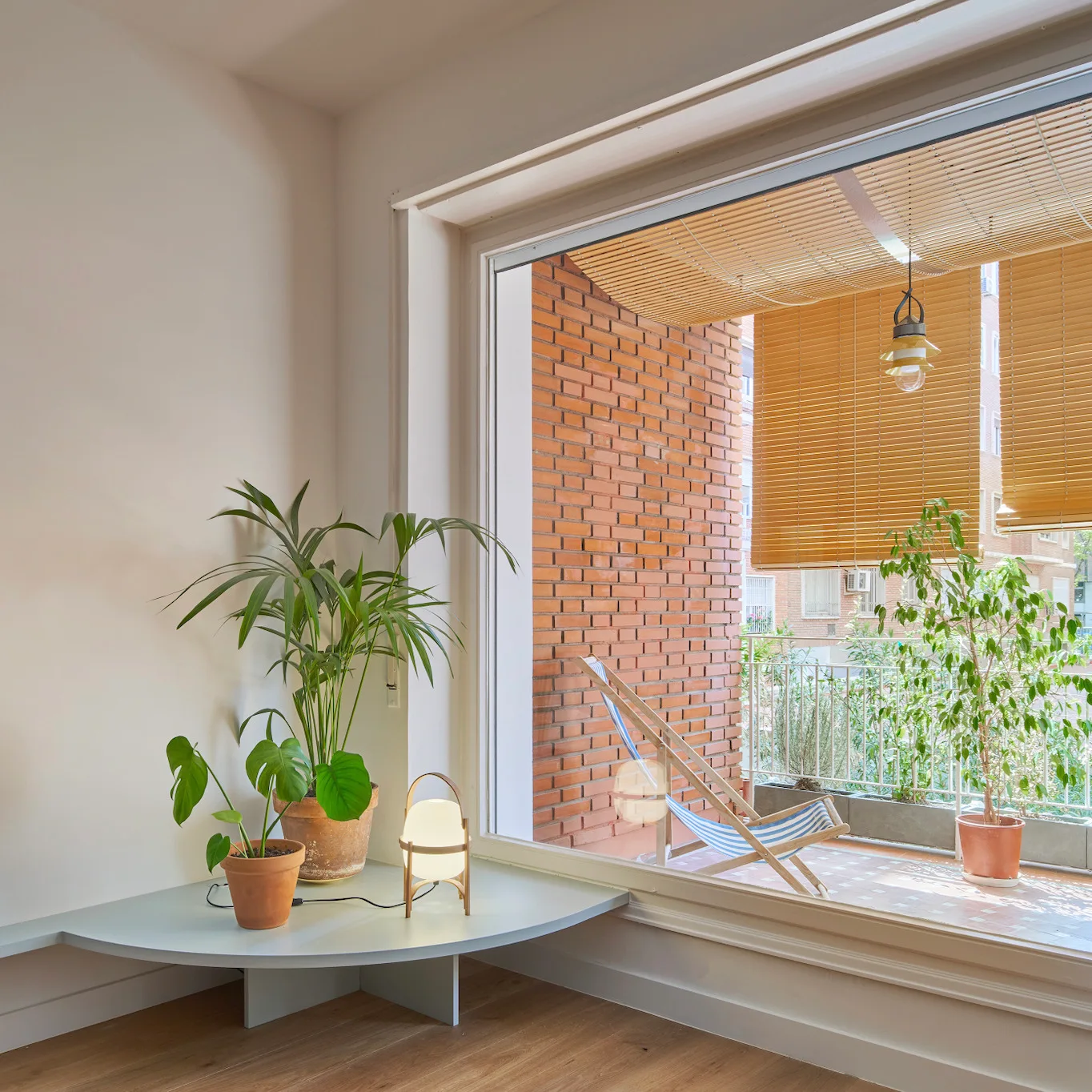 A low curved table with potted plants and a small lamp stands next to a large window showing a balcony with a brick wall and bamboo blinds.