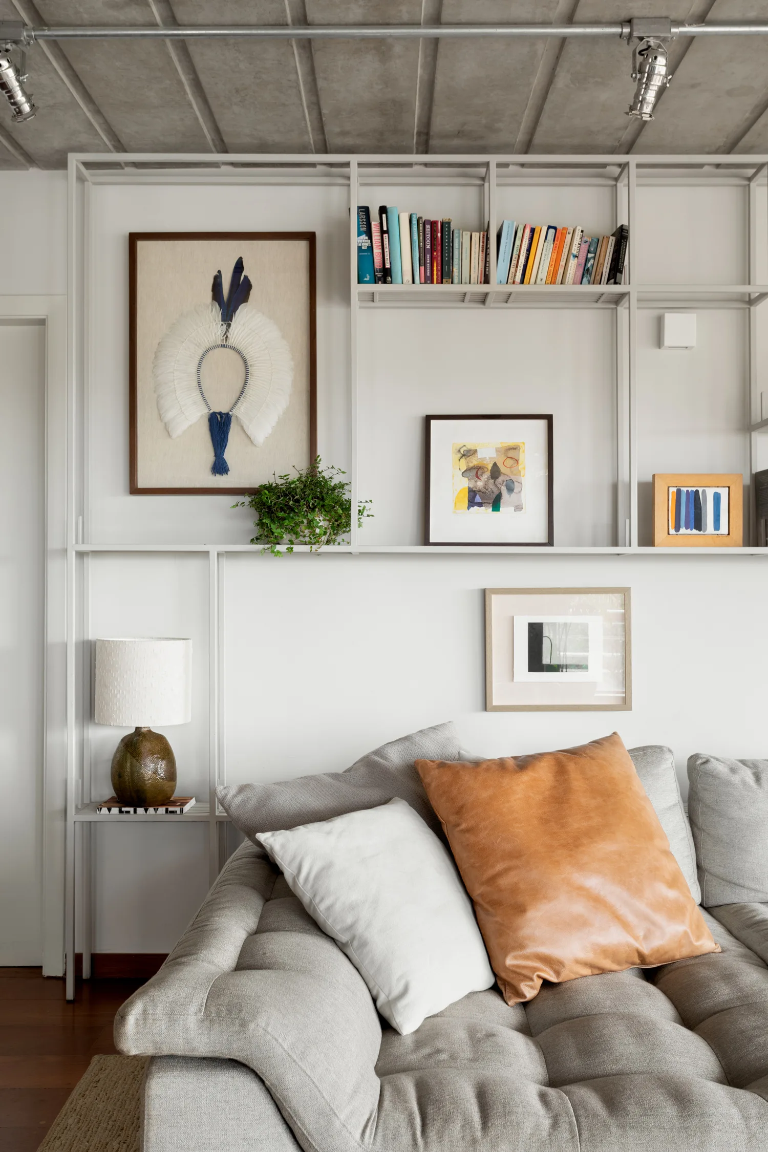 A tufted grey sofa with leather and white throw pillows sits below a tall metal grid shelving unit displaying books and framed artwork.