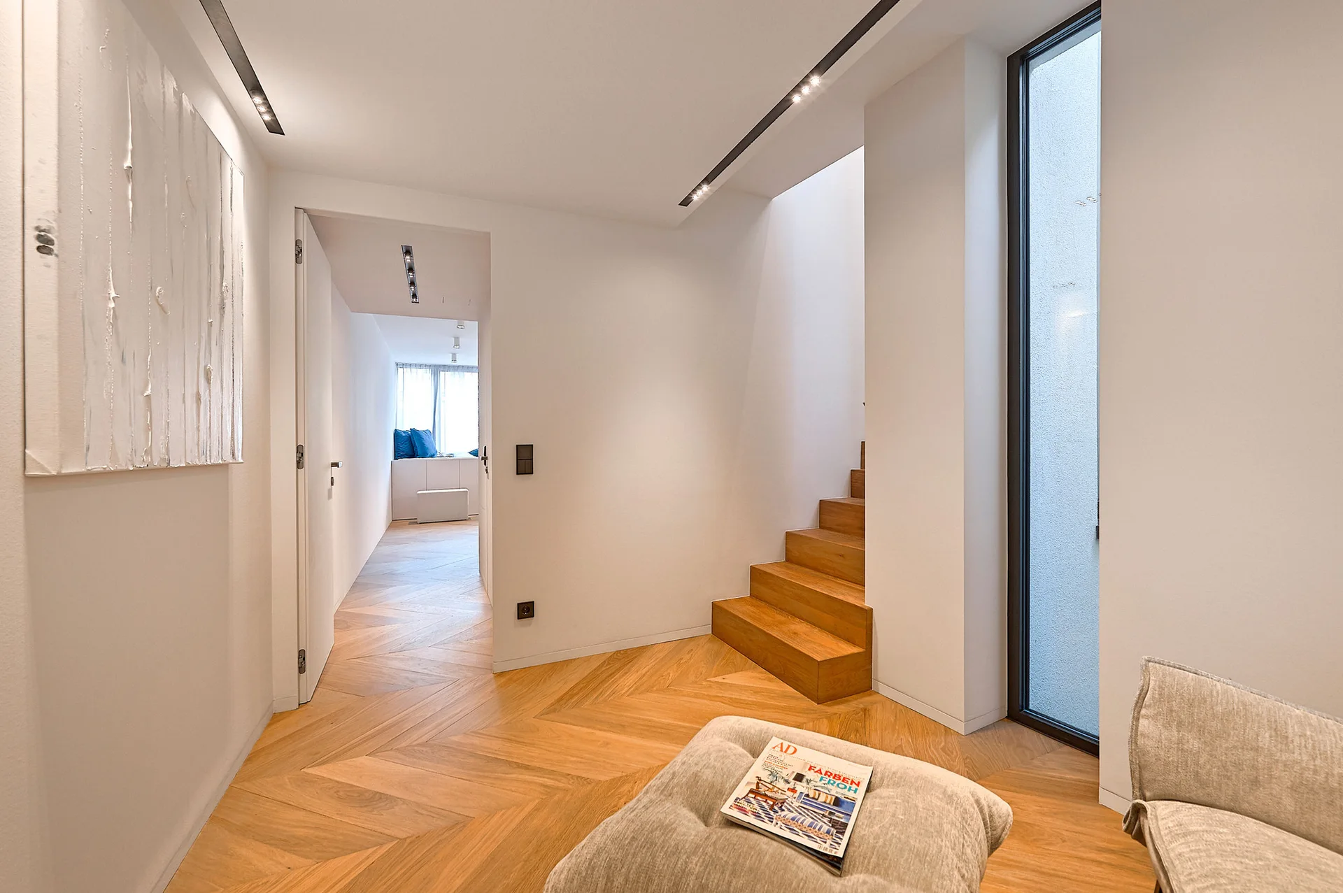 A minimalist hallway features light wood herringbone flooring, a wooden staircase, and white walls illuminated by recessed ceiling light tracks.