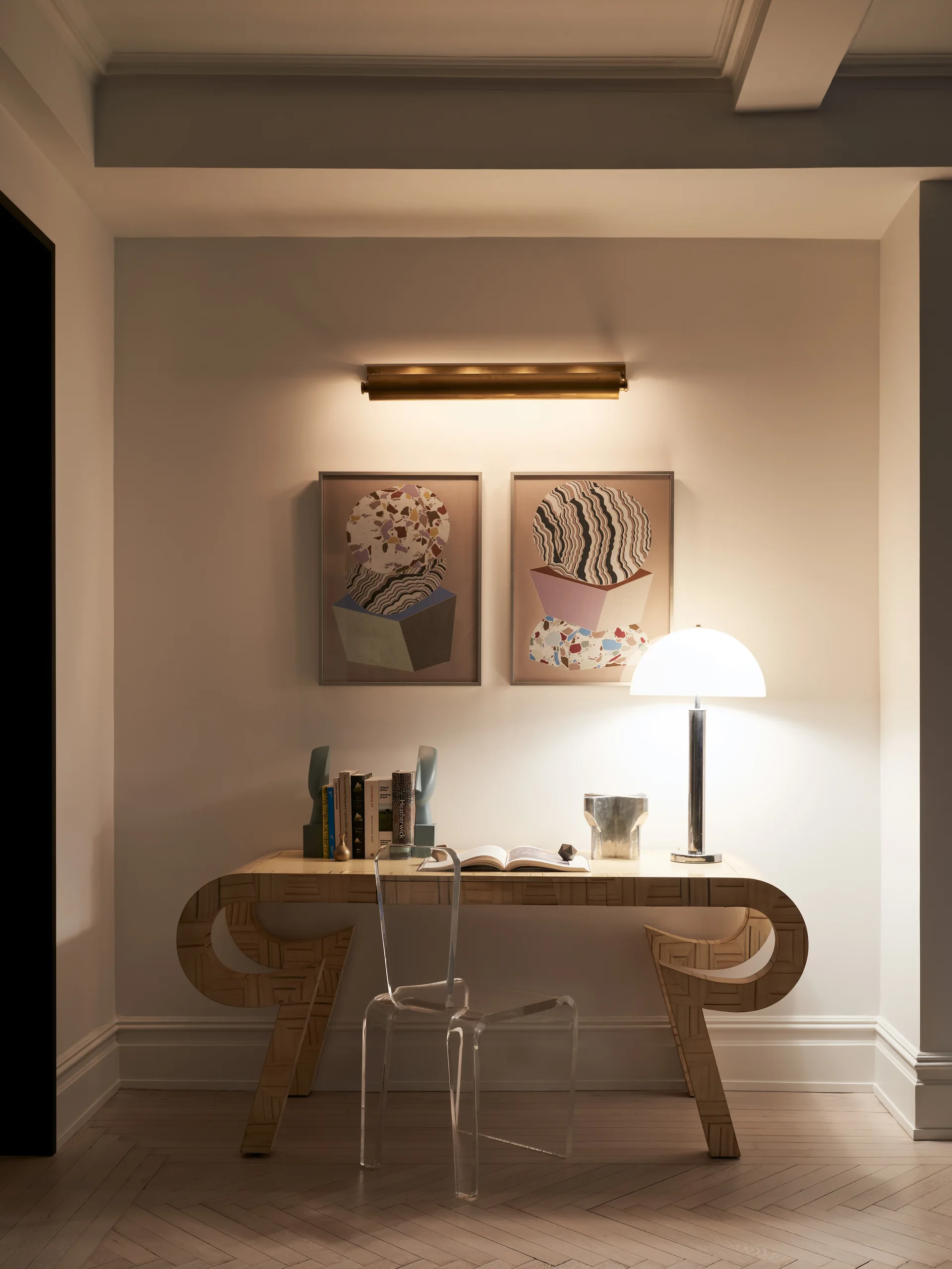 A light wood console desk with curved legs stands against a wall beneath two abstract framed prints, illuminated by a table lamp and brass sconce.