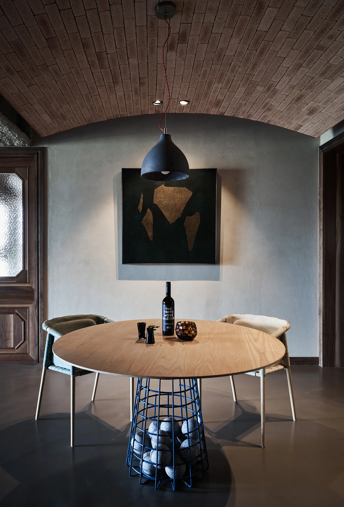 A round wooden dining table with a blue metal basket base sits under a vaulted brick ceiling and a black dome pendant light.
