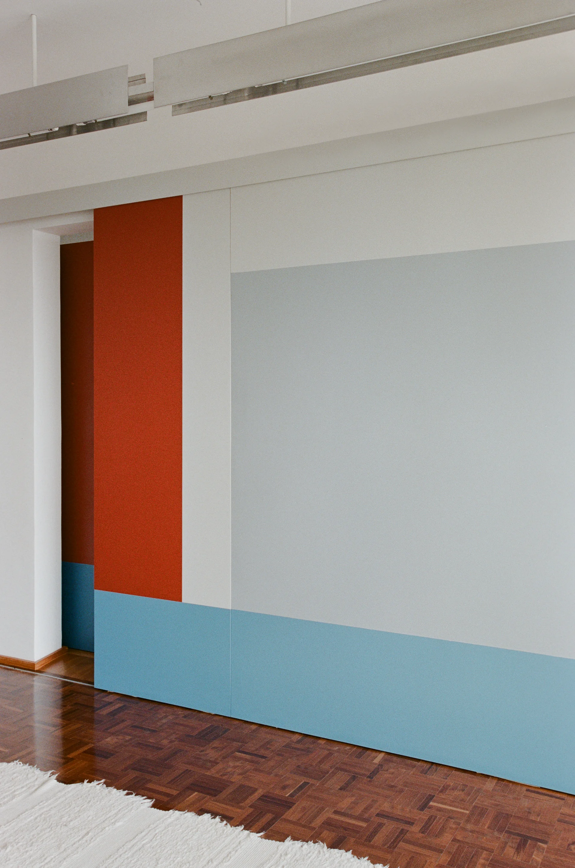 Matte geometric wall panels in red, blue, grey, and white sit above a dark wood parquet floor next to a white shag rug.