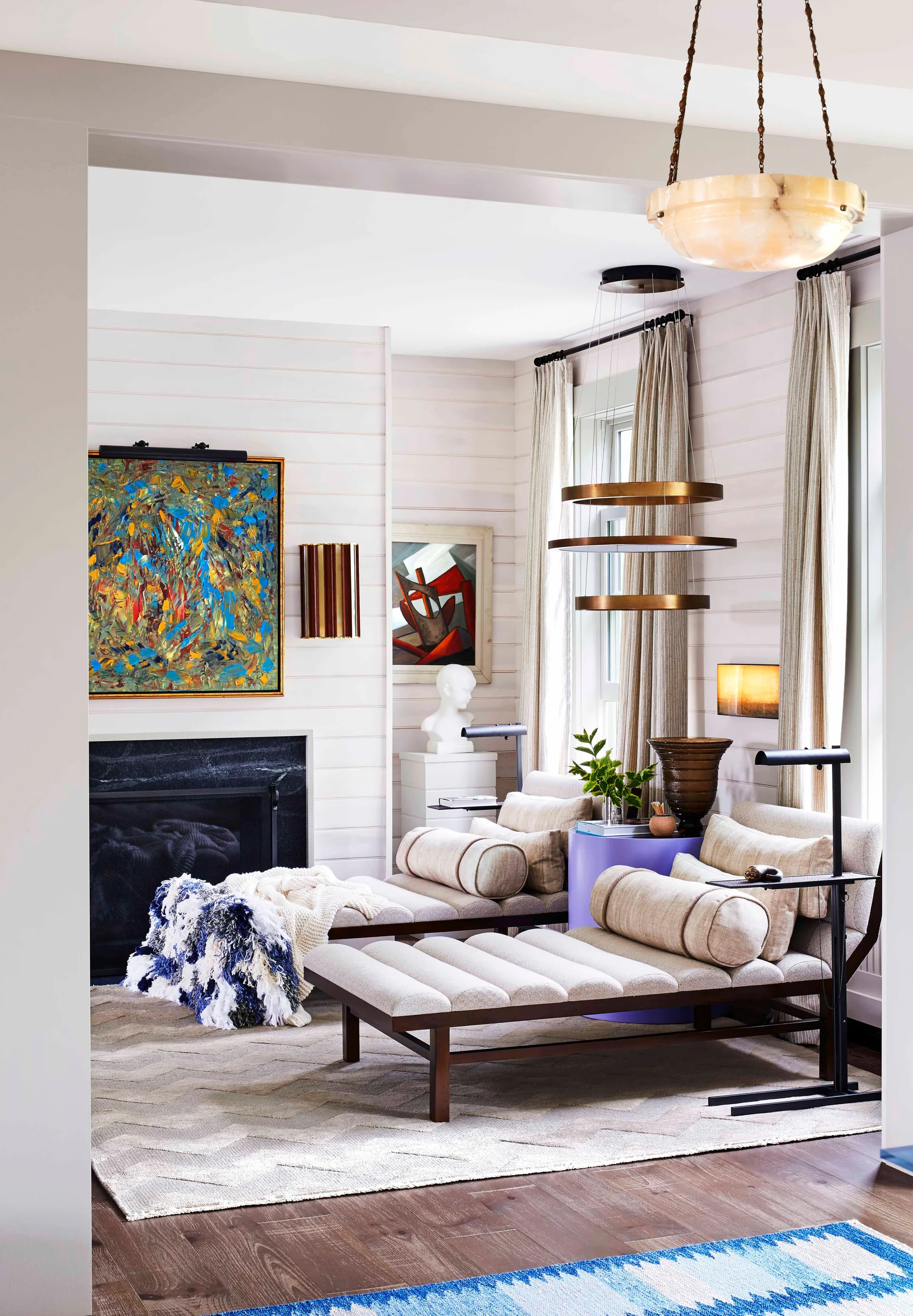 A sitting room features two upholstered daybeds separated by a purple side table on a chevron rug near a black fireplace and white wood-paneled walls.