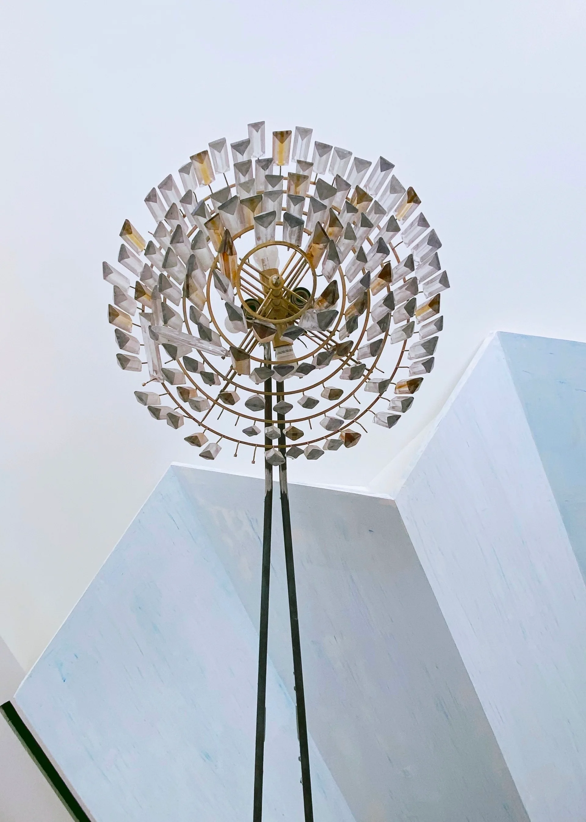 A low-angle view of a circular brass chandelier featuring concentric rings of clear geometric crystals against a plain ceiling.