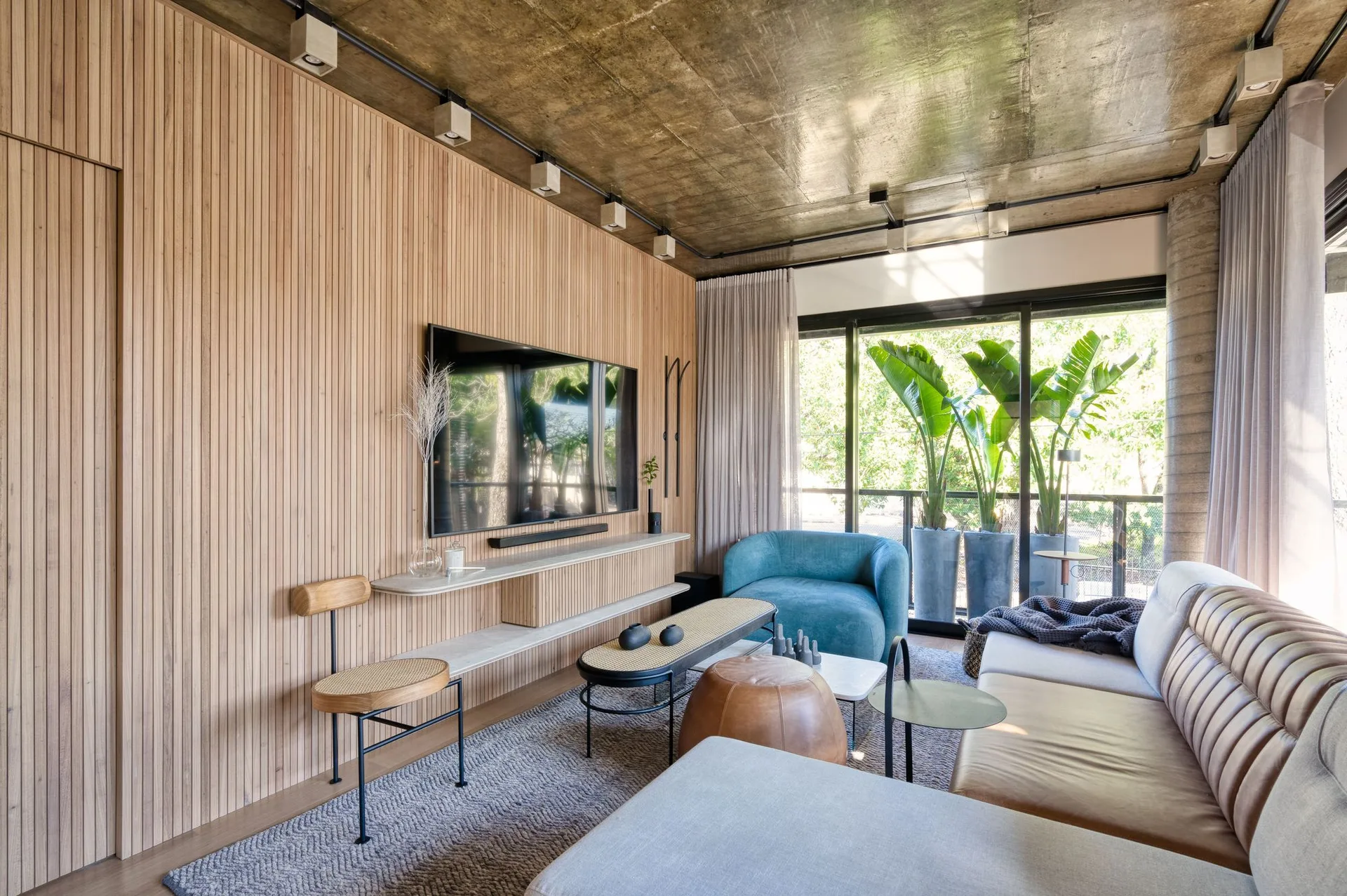 A modern living room features a vertical wood slatted wall, a mounted TV, a leather sofa, a blue armchair, and an exposed concrete ceiling.