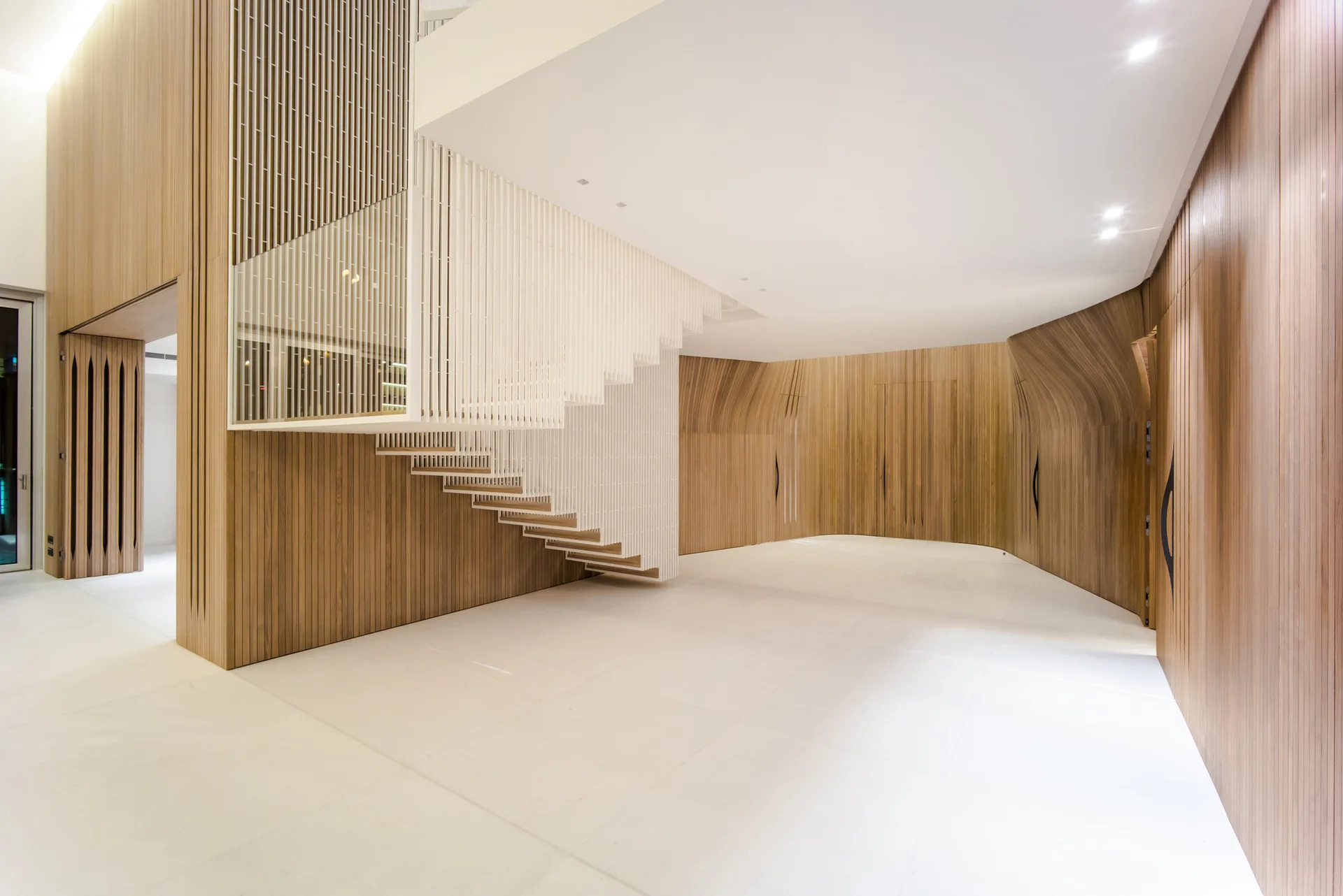 A minimalist entryway showcases a white suspended staircase with vertical slats alongside curved wood-paneled walls and clean white floors.