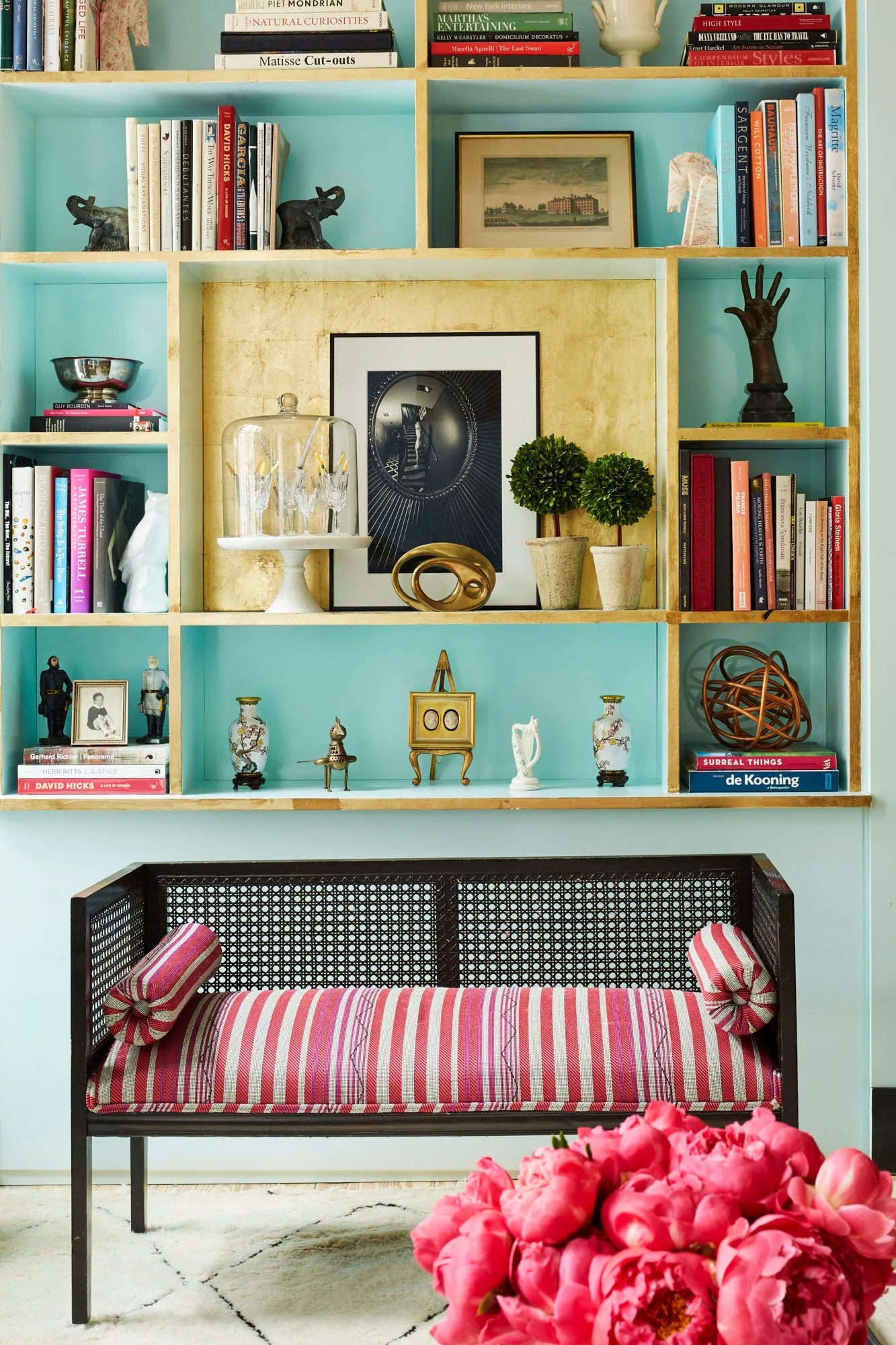A black cane bench with red and white striped upholstery sits in front of a bright turquoise built-in shelving unit filled with books and art objects.