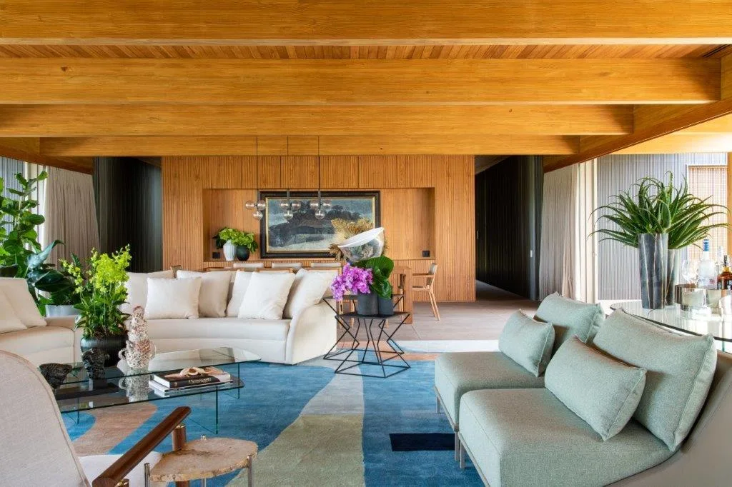 A contemporary living room featuring exposed wood ceiling beams, a slatted timber accent wall, and a large blue patterned rug beneath white and sage sofas.