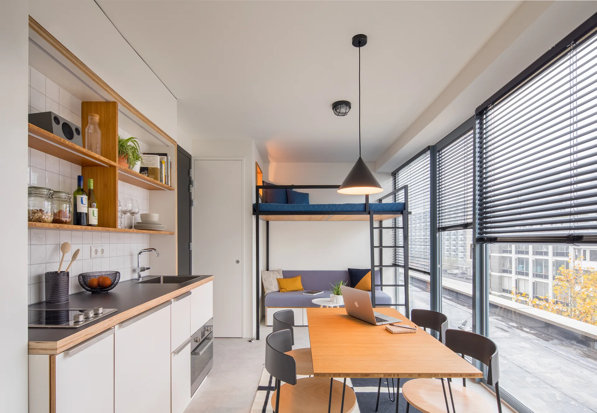 A compact studio apartment features a light wood dining table, a white kitchen with open shelving, and a loft bed over a grey sofa.