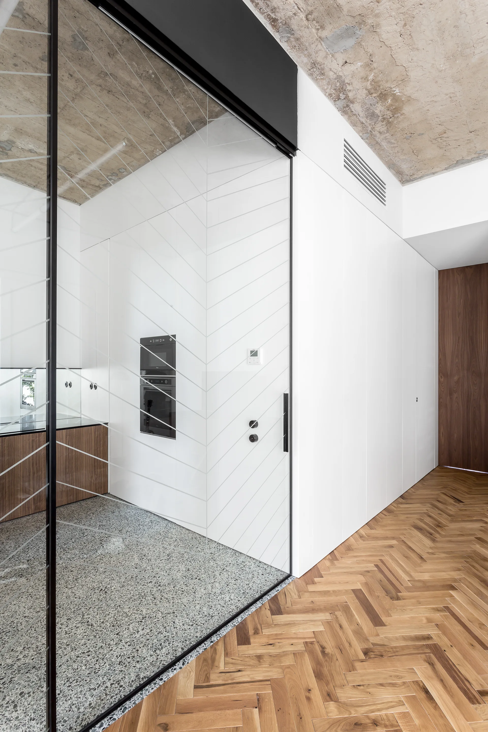 A modern kitchen with terrazzo flooring is separated by a glass sliding partition from a hallway featuring herringbone wood flooring.