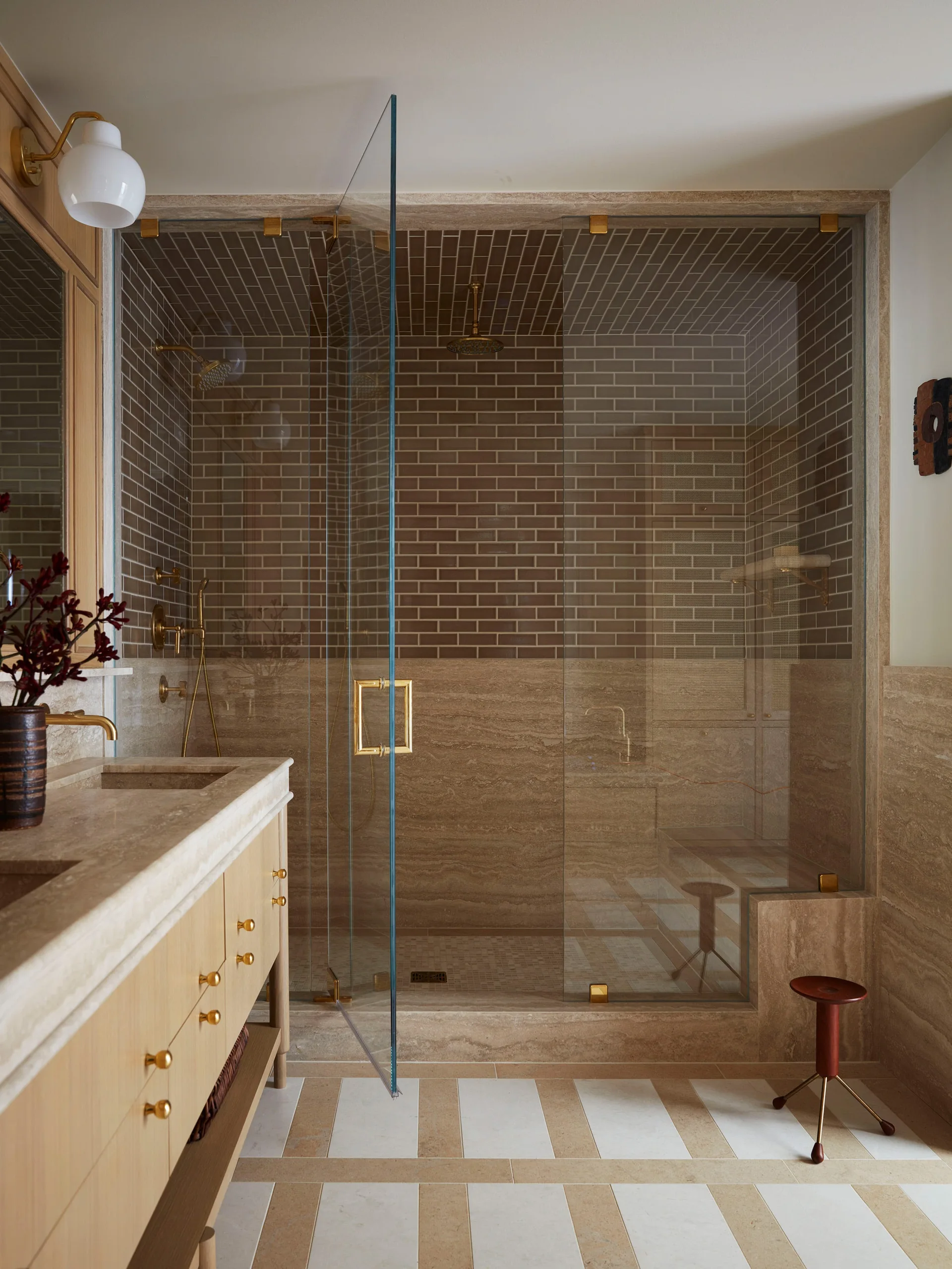 A modern bathroom features a light wood vanity, a glass walk-in shower with brown subway tile, and a striped tile floor.