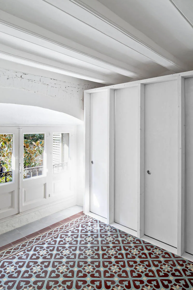 A minimalist white room with patterned burgundy and blue floor tiles, a built-in closet, and a beamed ceiling.