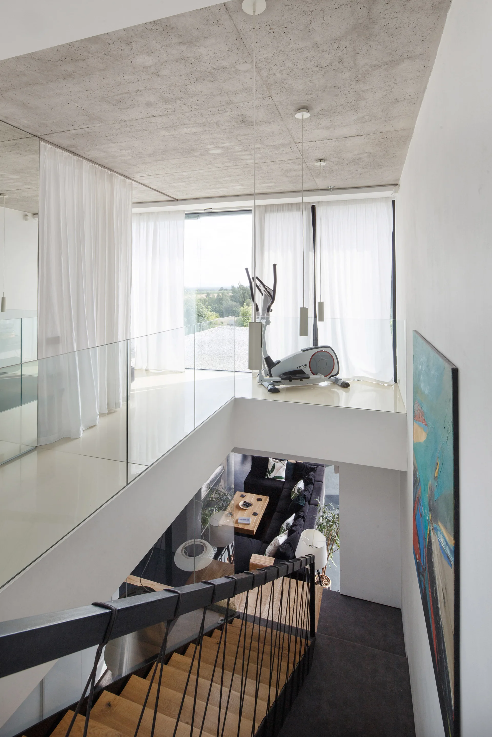 A view down a wooden staircase from a modern white landing with an elliptical machine, glass railings, and an exposed concrete ceiling.