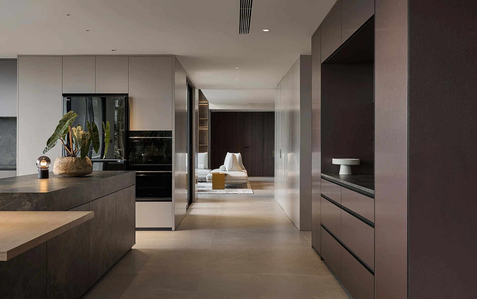A modern minimalist kitchen featuring matte taupe cabinetry, a grey stone island with a potted plant, and a hallway leading to a living area.